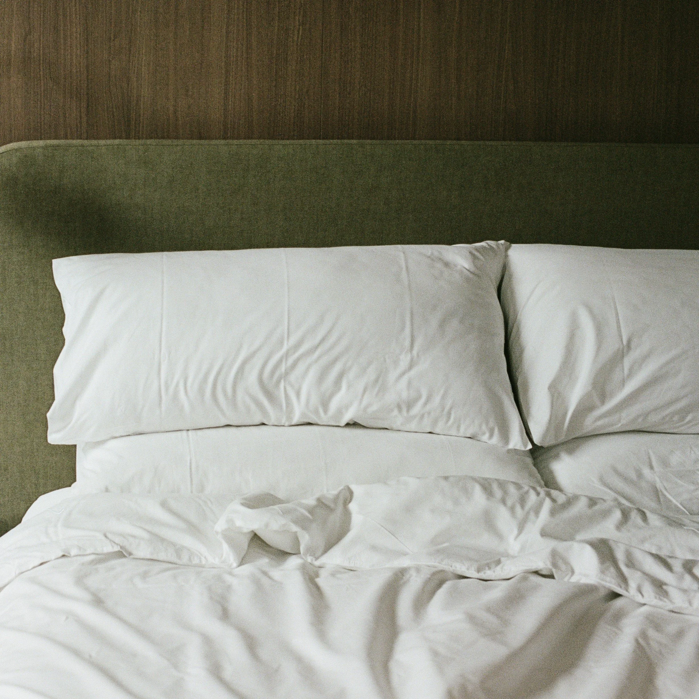 A photograph of a white pillow on a bed with a crumpled white sheet and a green headboard against a brown wooden wall.