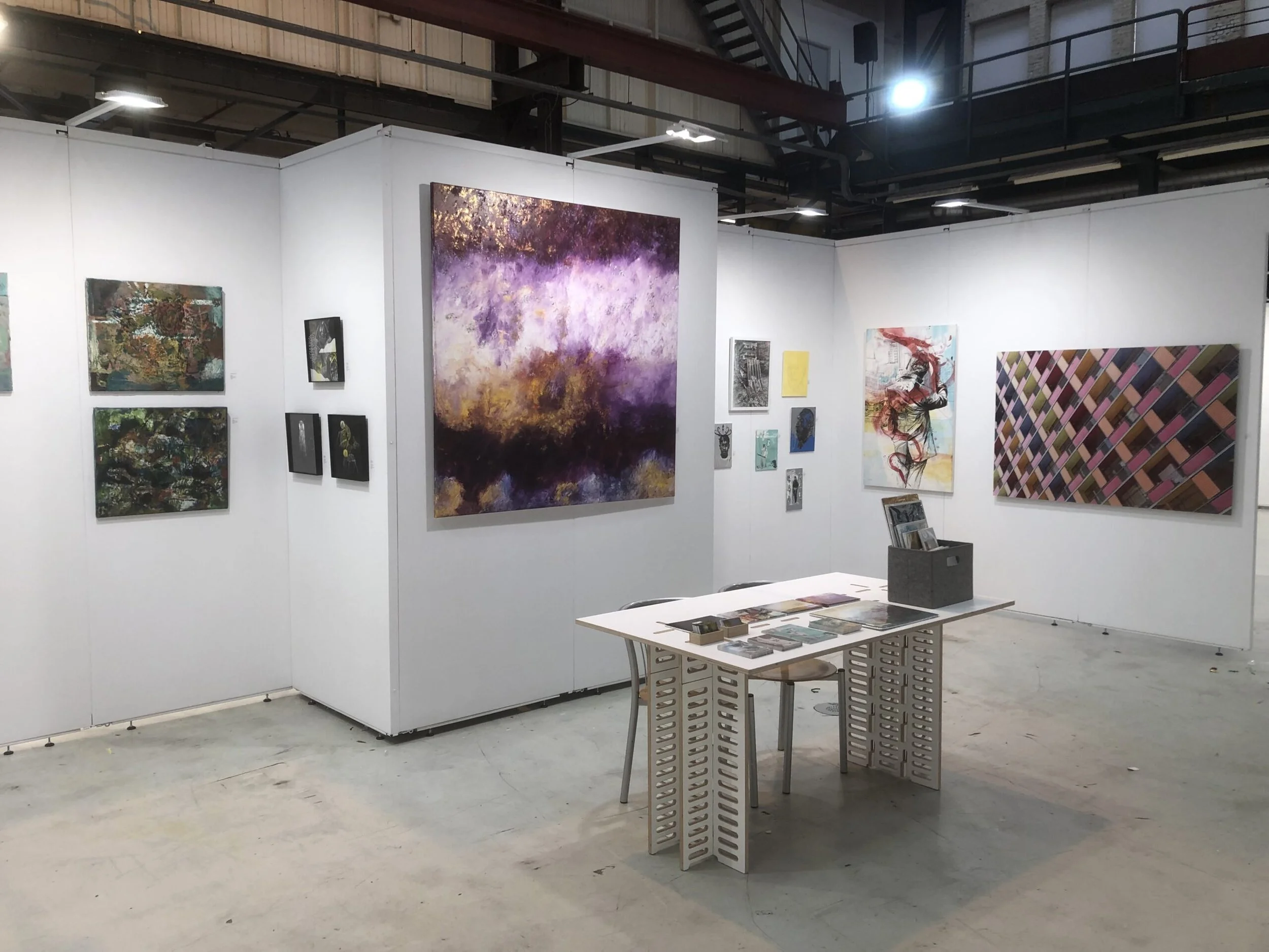 Art gallery with white display walls showcasing various paintings and drawings, a table with pamphlets, in an industrial-style space with exposed ceiling beams and lighting.