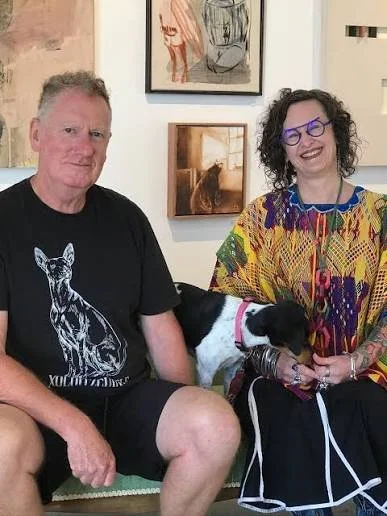 A man and woman sitting on a bench indoors with a black and white dog between them. The man is wearing a black t-shirt with a dog graphic, and the woman is wearing colorful clothing and purple glasses, smiling. There are framed artworks on the wall behind them.