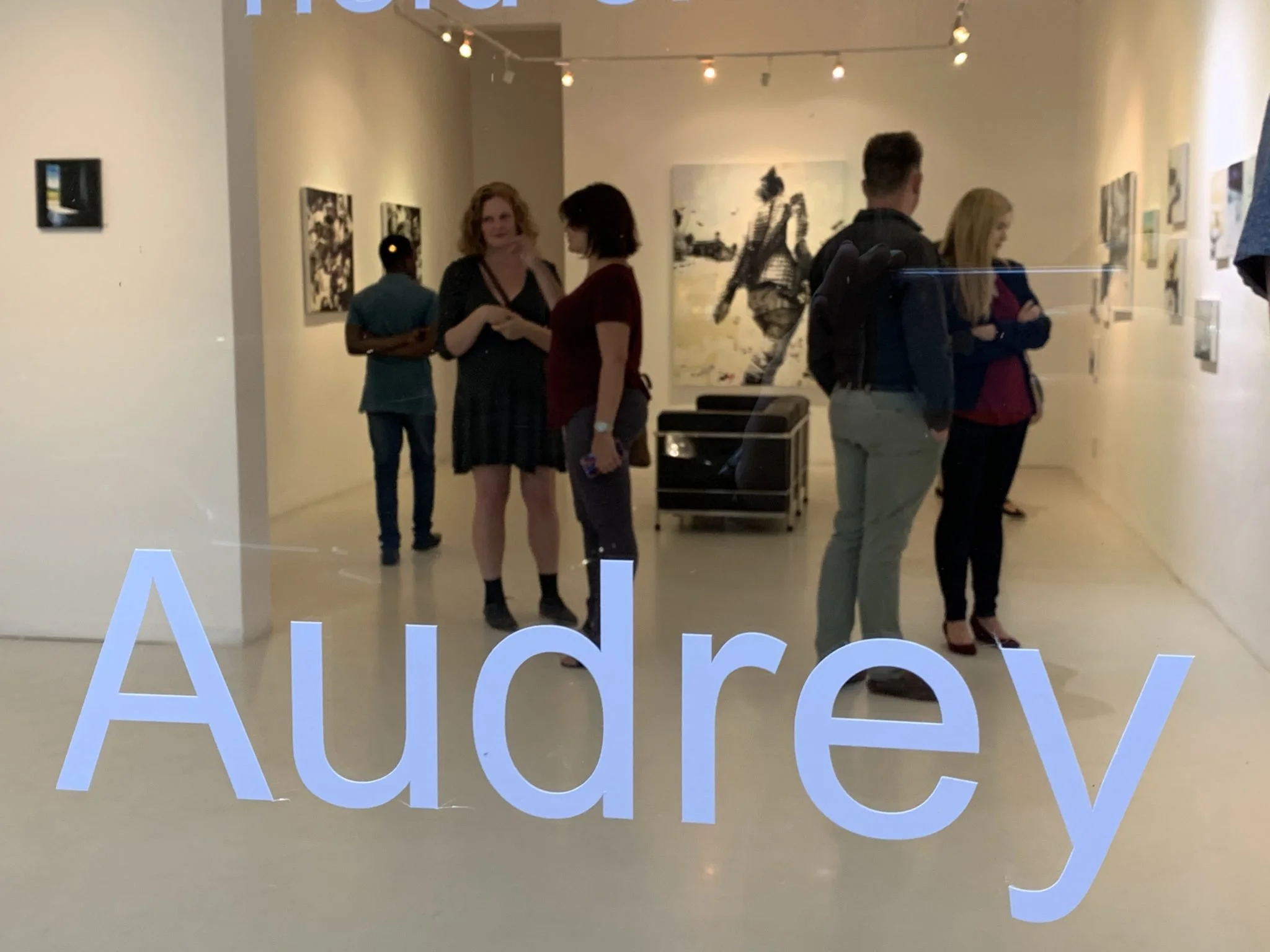 People viewing artwork in an art gallery with large white letters spelling 'AUDIO' on the floor in the foreground.