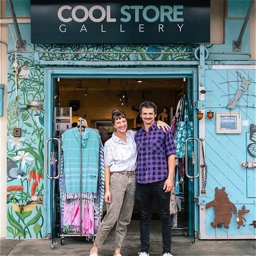 Two people standing outside the entrance of Cool Store Gallery. The woman is wearing a white blouse and beige pants, and the man is wearing a purple checkered shirt and dark pants. The entrance is decorated with colorful murals and display racks with clothing.