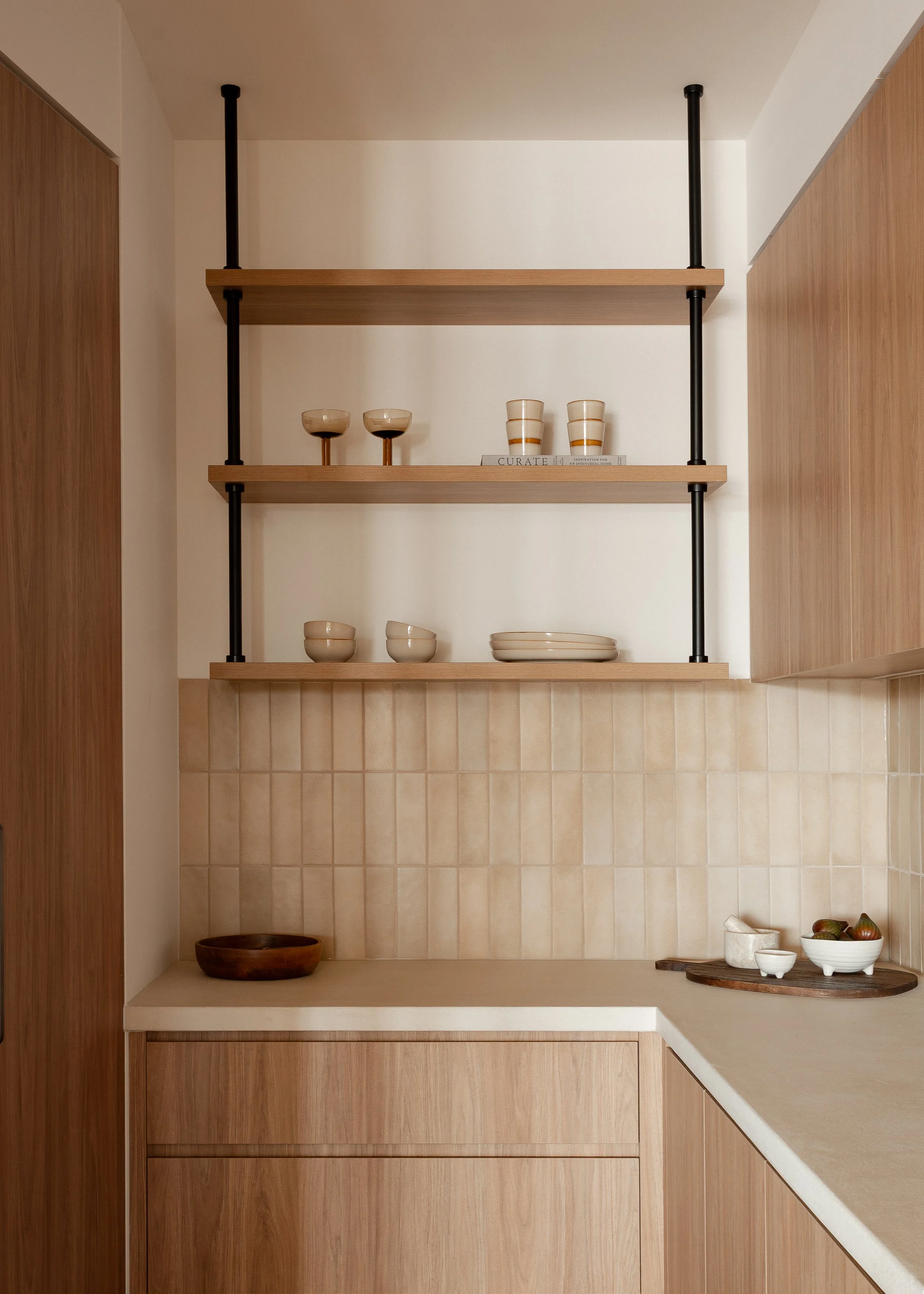 Open shelves kitchen walk in pantry with three shelves holding ceramic dishes, bowls, glasses, and a wooden bowl, with a tiled backsplash and wooden cabinetry.