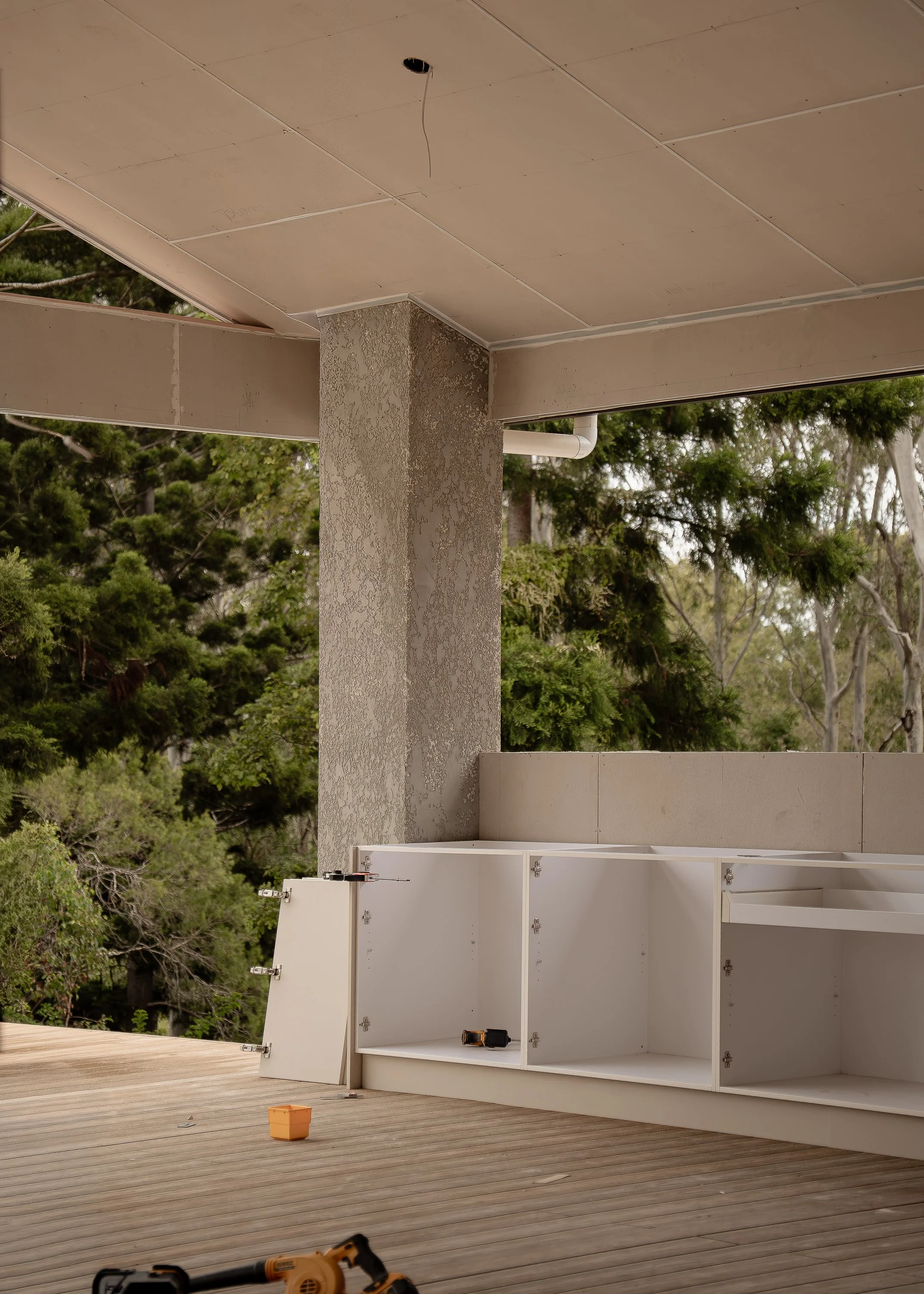 Under construction or renovation scene showing a partially assembled white cabinet on a wooden deck with tools and a small orange bucket, surrounded by trees and outdoor environment.