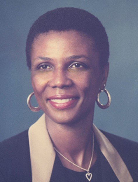 Portrait of a woman with short hair, wearing hoop earrings, a heart-shaped necklace, and a black blazer with a beige collar, smiling against a plain background.