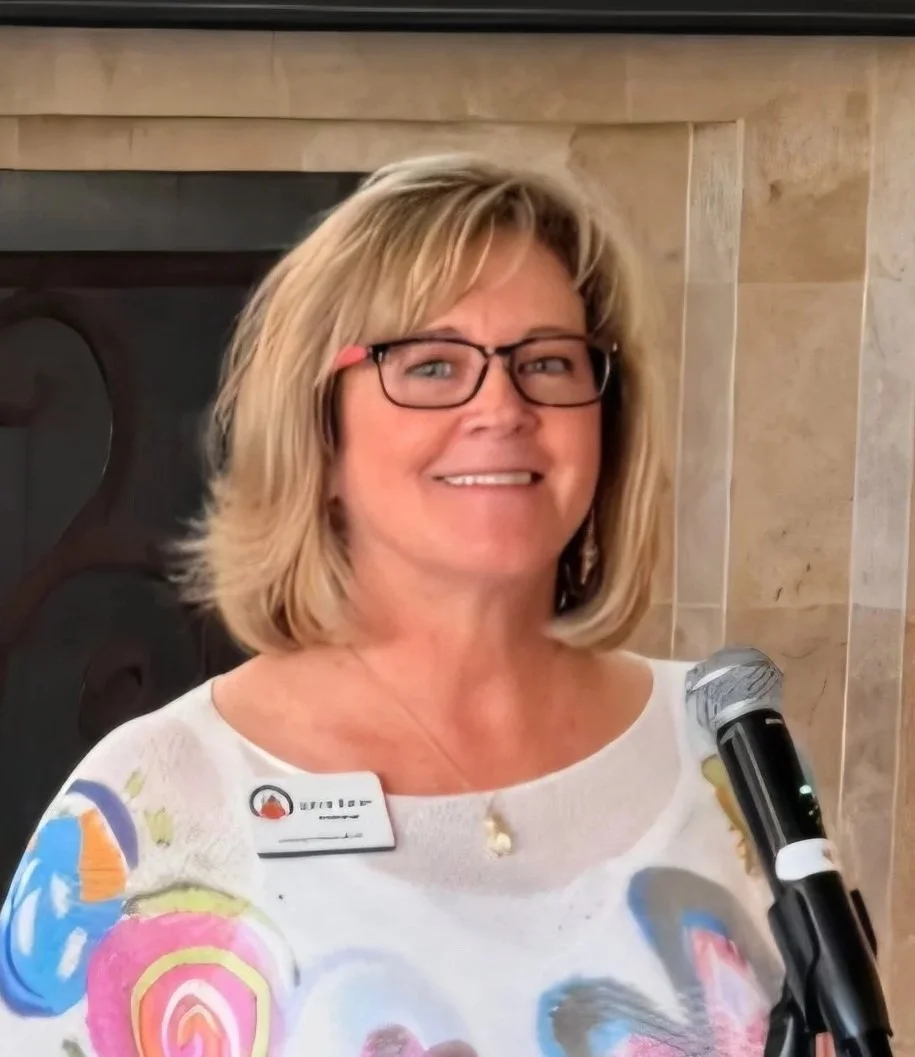 A woman with glasses and shoulder-length blonde hair smiling, holding a microphone, standing in front of a fireplace with a marble tile wall. She is wearing a white top with colorful abstract patterns and a name tag.