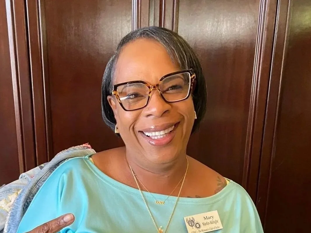 A woman with short black hair, wearing glasses, a turquoise blouse, and a gold necklace, smiling and pointing at her name badge that reads Mary.