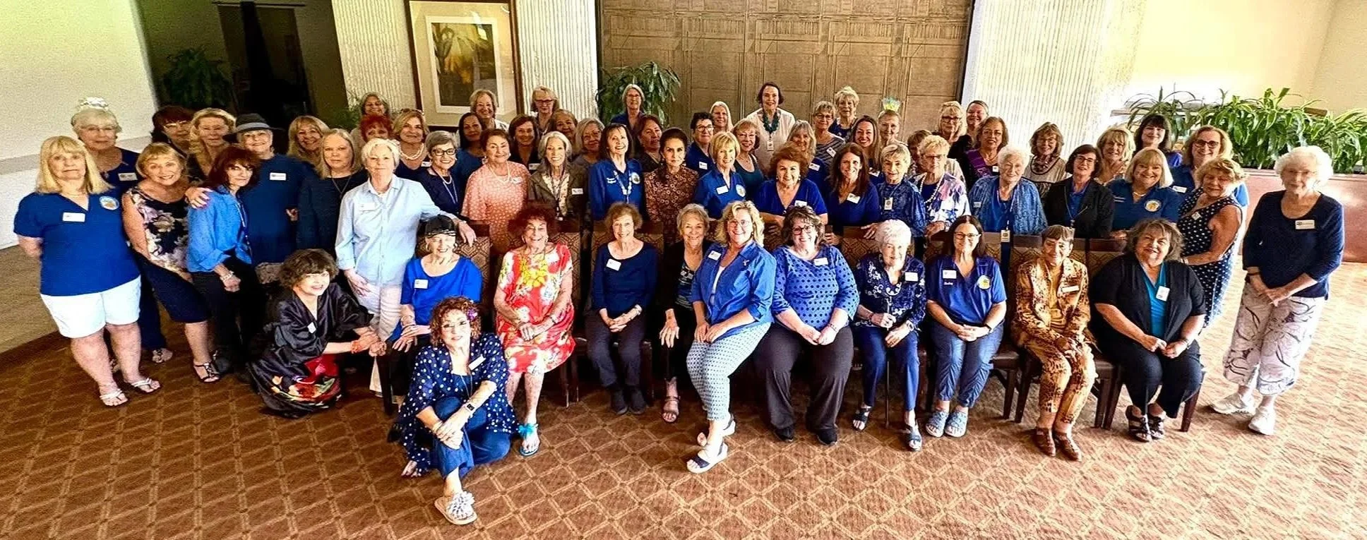 A large group of women gathered indoors for a group photo, many wearing blue tops, with some seated and others standing, in a room with artwork, plants, and vertical blinds.