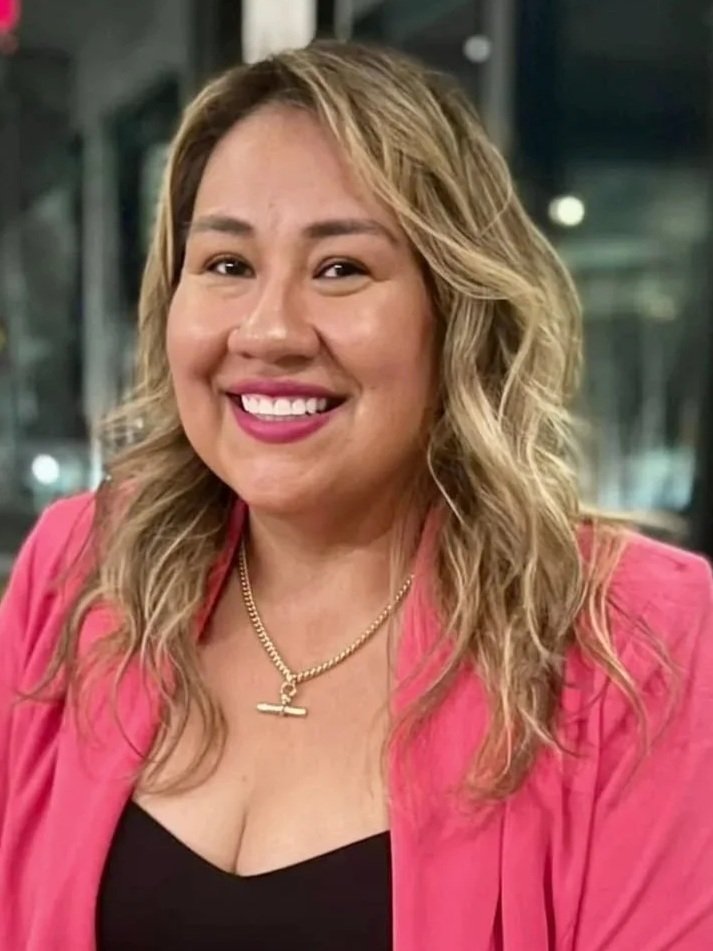 A woman with blonde, wavy hair smiling, wearing a pink blazer over a black top, and a gold necklace with a pendant.