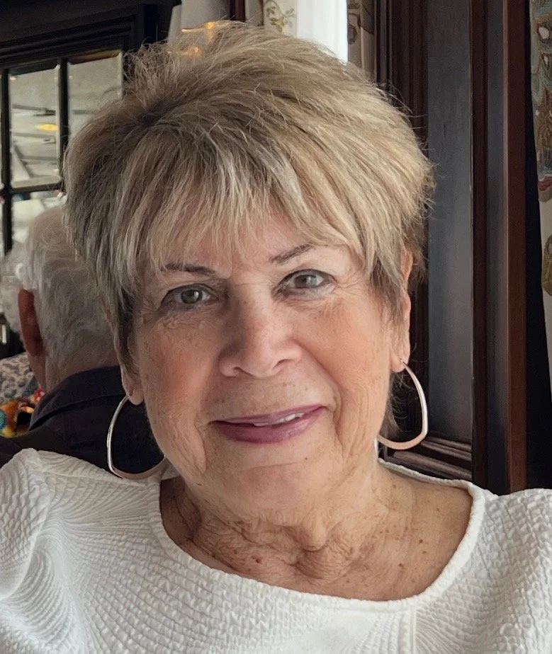 Close-up of an elderly woman with short, light blonde hair, wearing large hoop earrings and a white textured top, sitting in a restaurant or cafe.