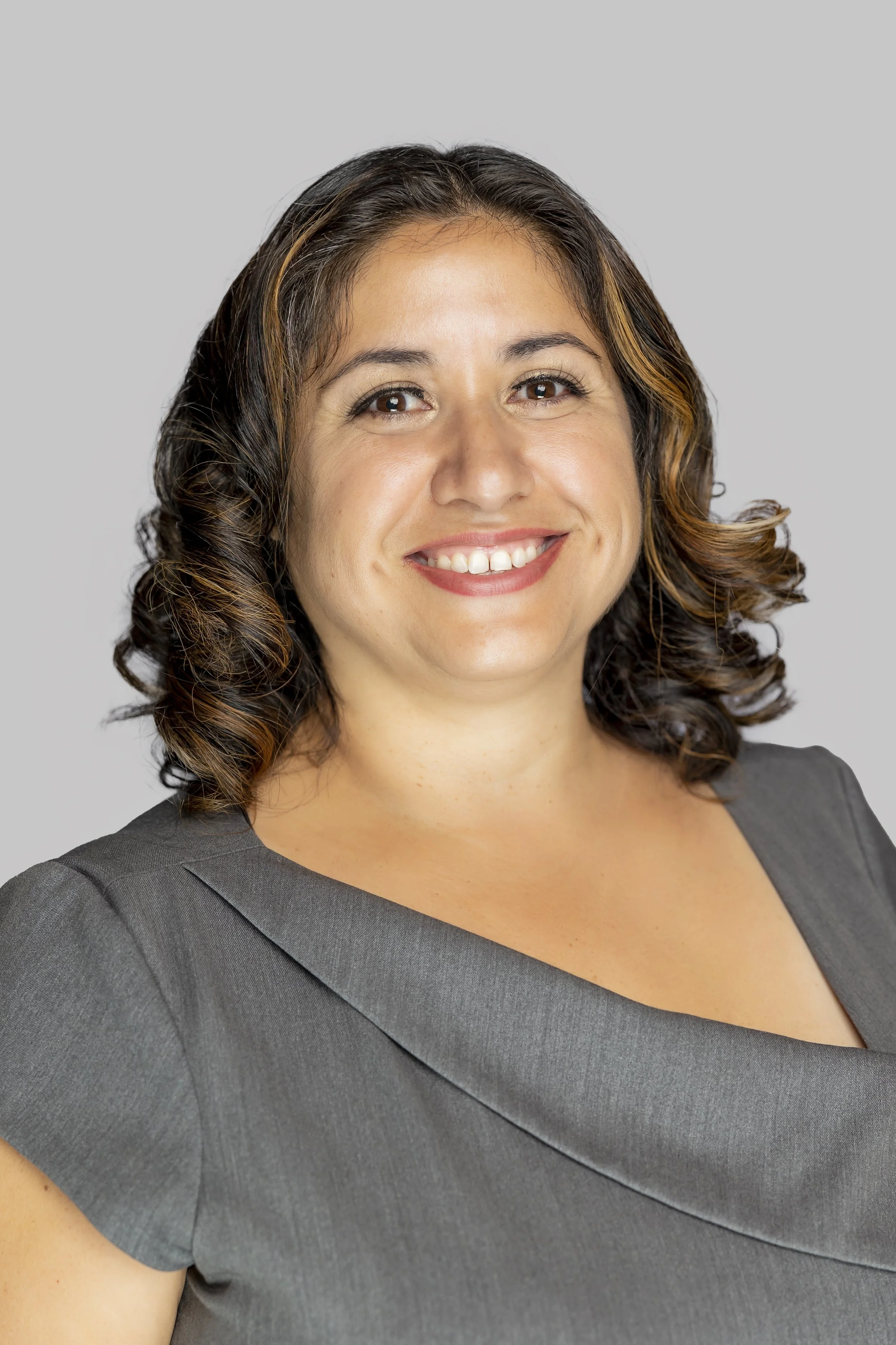 Close-up of a woman with shoulder-length curly brown hair, wearing a dark gray dress, smiling with white teeth, standing against a plain light gray background.
