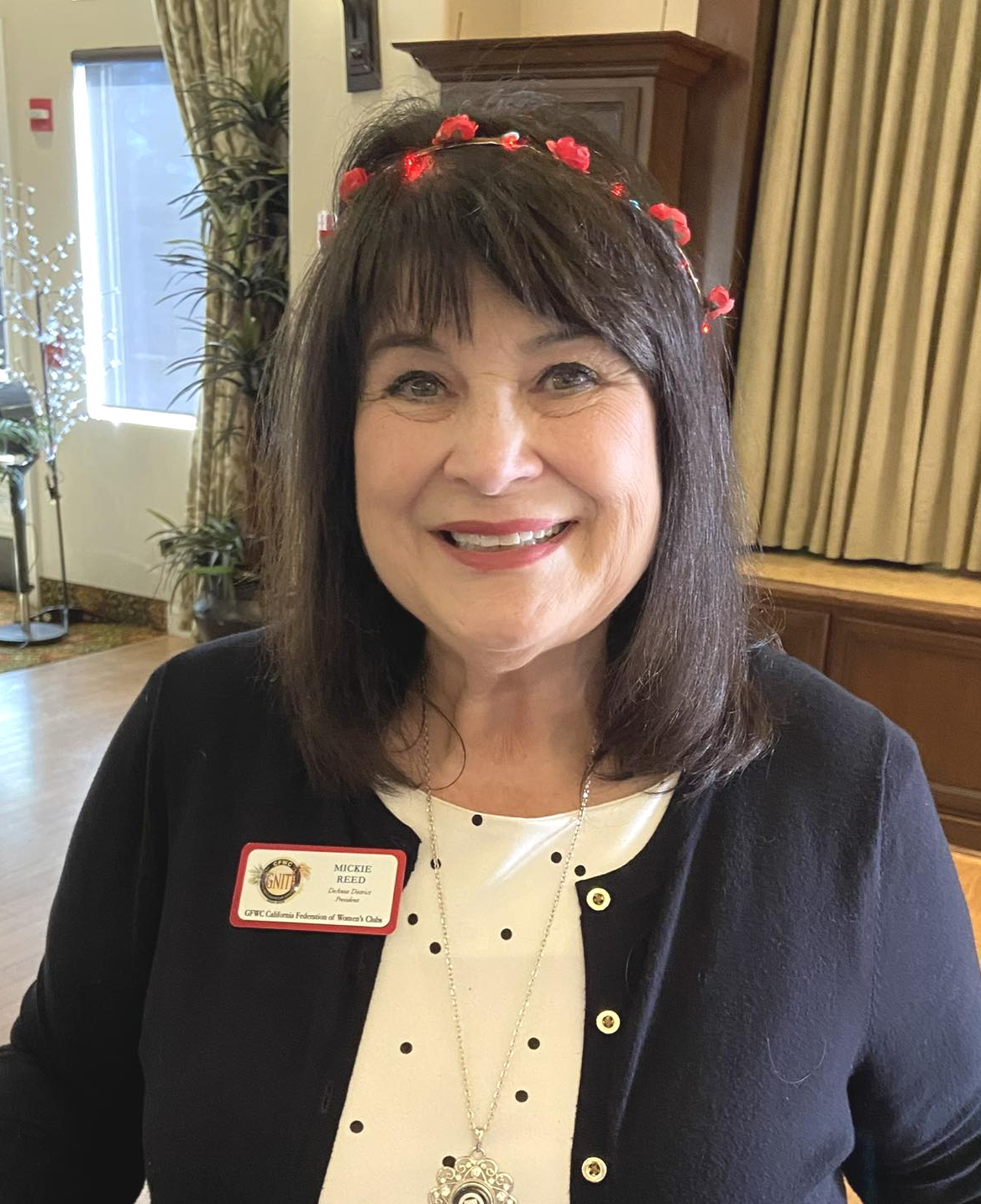 A smiling woman with shoulder-length dark hair, wearing a black jacket over a white polka dot blouse, a floral headband with red flowers, and a name badge that reads 'Mickie Reed, District Director, GFW California Federation of Women's Clubs.'