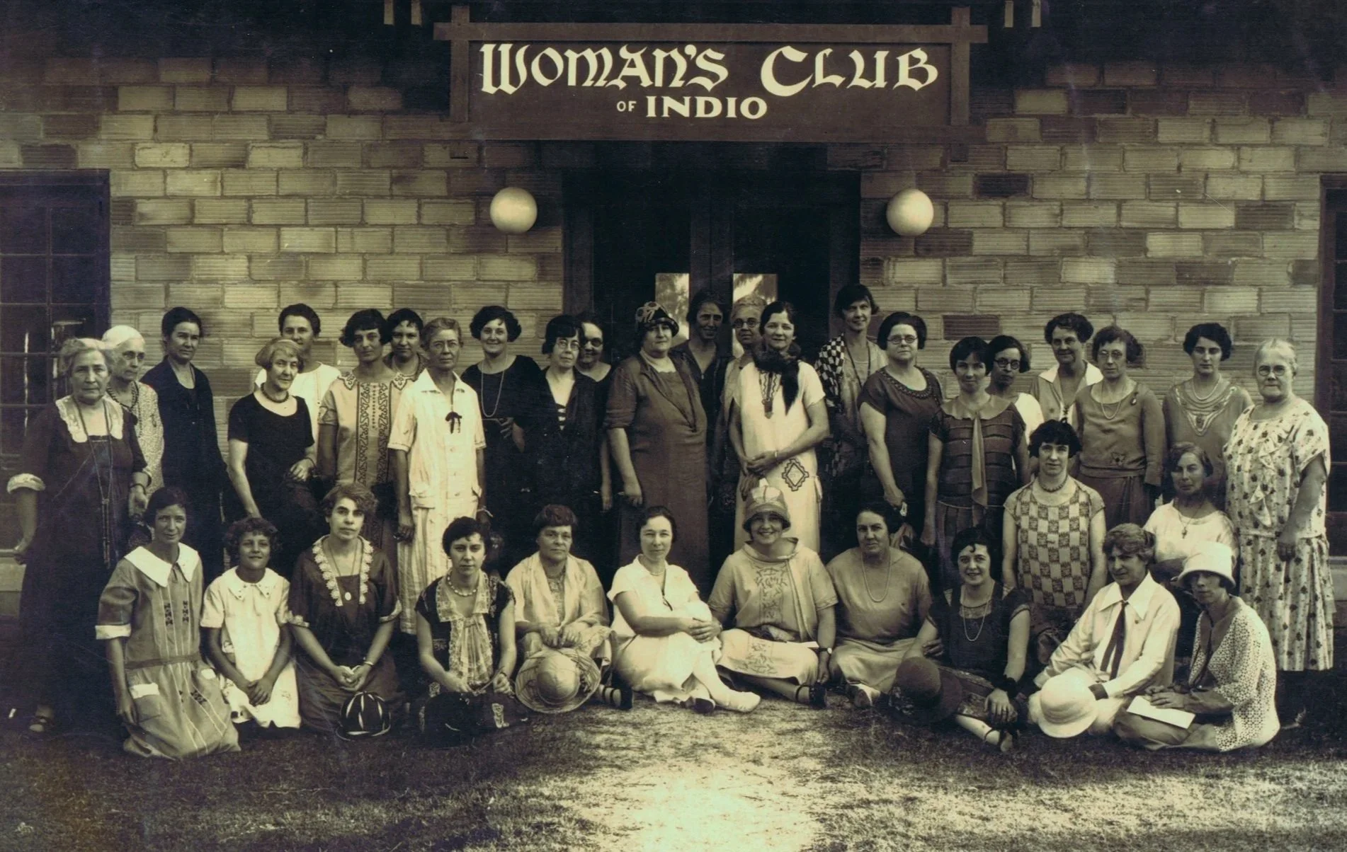 A group of women posed for a photograph in front of a brick building with a sign that reads "Woman's Club of Indio." The women are wearing vintage clothing and are original club members in this 1925 photo.