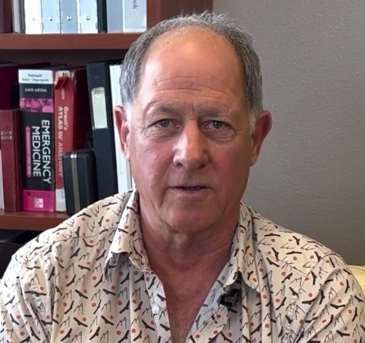 A middle-aged man with short brown hair and blue eyes is sitting in an office, wearing a patterned button-up shirt. In the background, there is a bookshelf with medical textbooks, including one titled "Emergency Medicine."