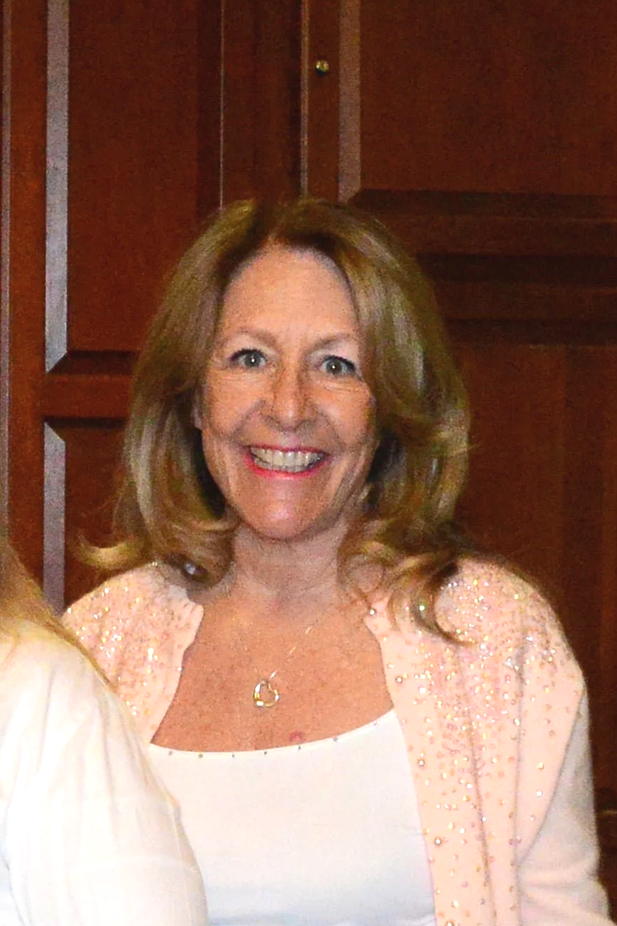 A woman with shoulder-length light brown hair, smiling and wearing a white top with a pink, sparkly jacket, standing in front of a wooden wall.