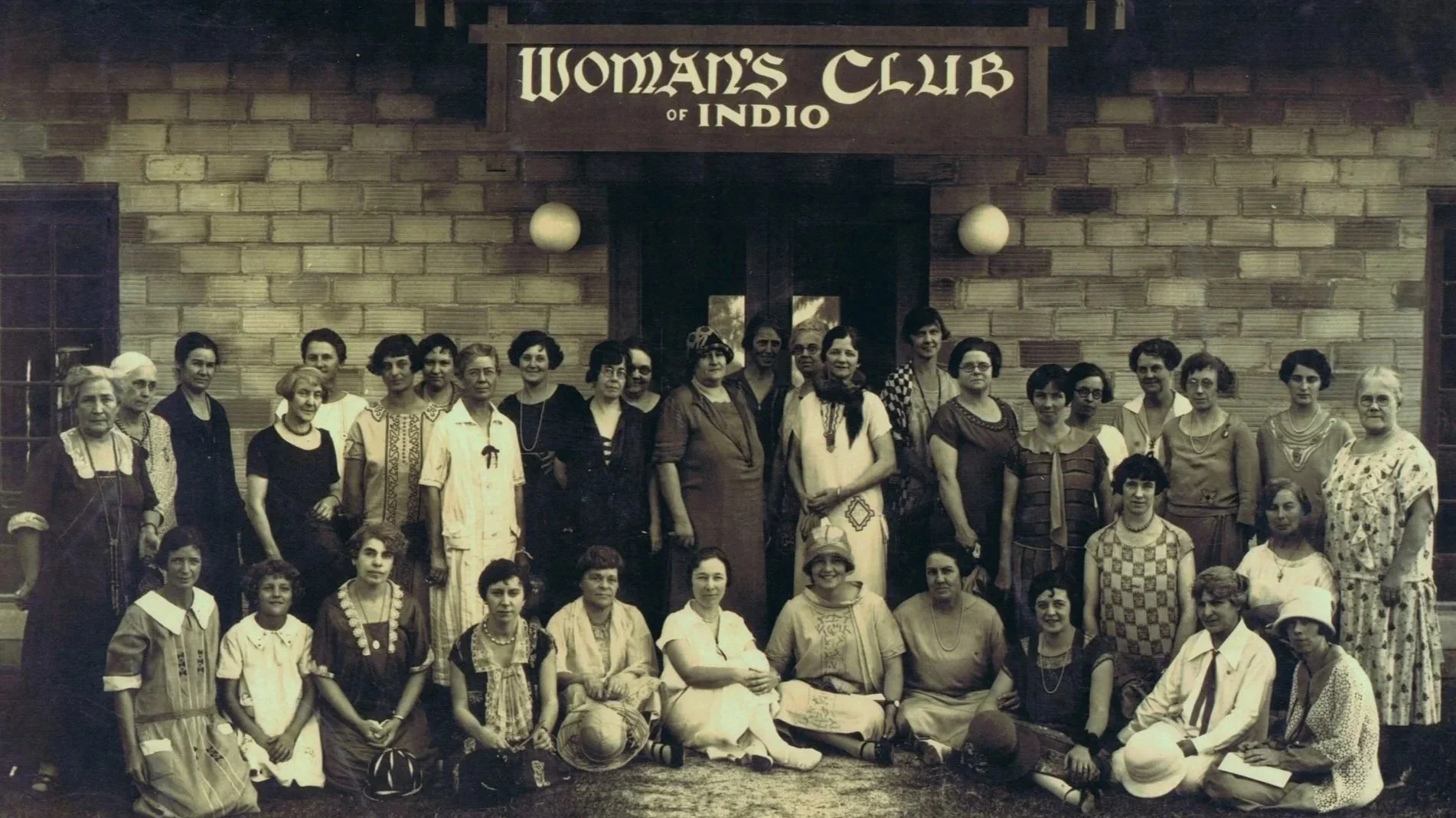 Group of women standing and sitting in front of a sign that reads 'Woman's Club of Indio', in front of a brick building.