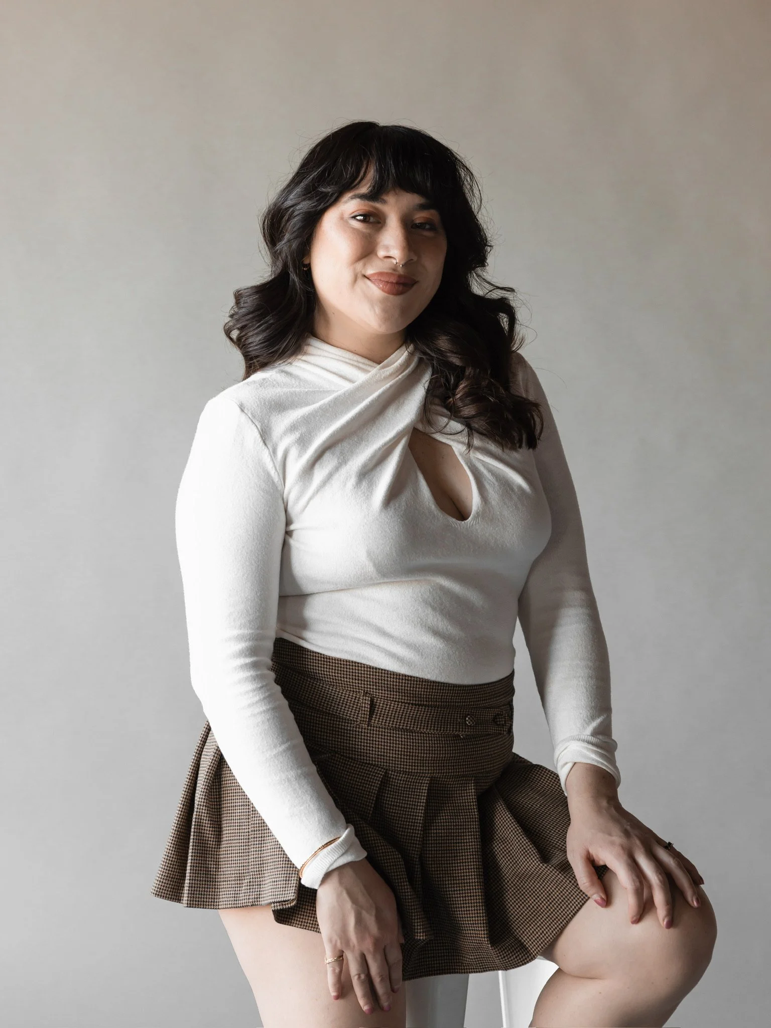 A woman sitting on a chair with a neutral background, wearing a beige top and a brown plaid skirt, with her hand resting on her knee.