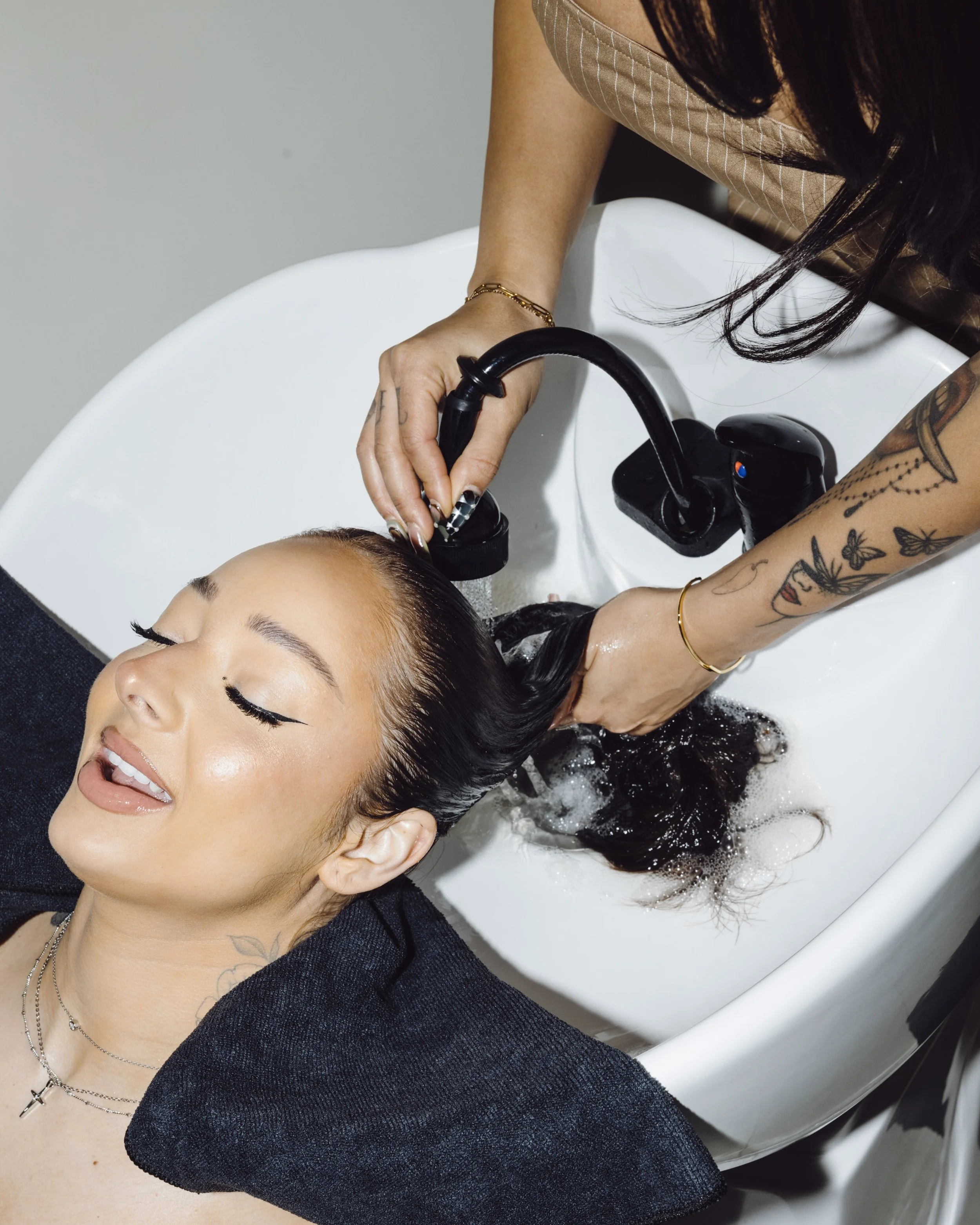 Person with dark hair getting their hair washed at a salon, lying back with eyes closed, in a white wash basin, with tattoos visible on arm and motivational necklace visible.