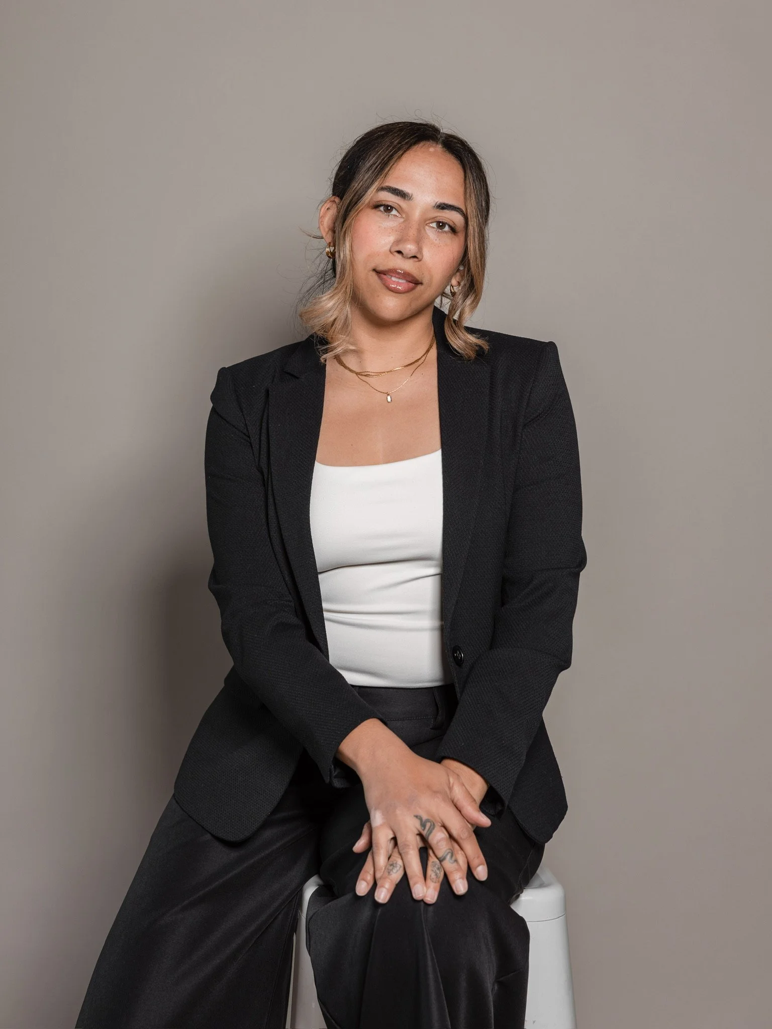 A woman with shoulder-length hair, wearing a black blazer, white top, and jewelry, sitting on a white stool against a plain gray background.