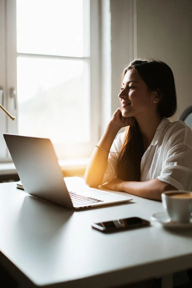 A woman sitting at a white desk in front of a laptop and a smartphone, in a bright room with natural light coming through a window.