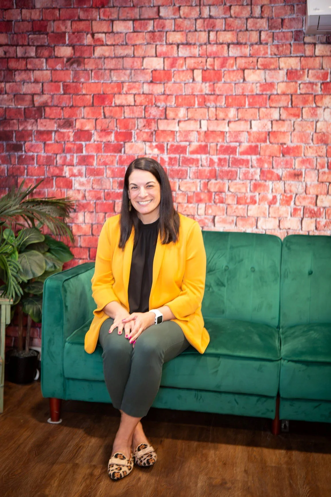 A woman with dark hair, wearing a yellow blazer, black top, green pants, and leopard-print shoes, sitting on a green velvet couch against a red brick wall background, smiling and holding her hands in her lap.