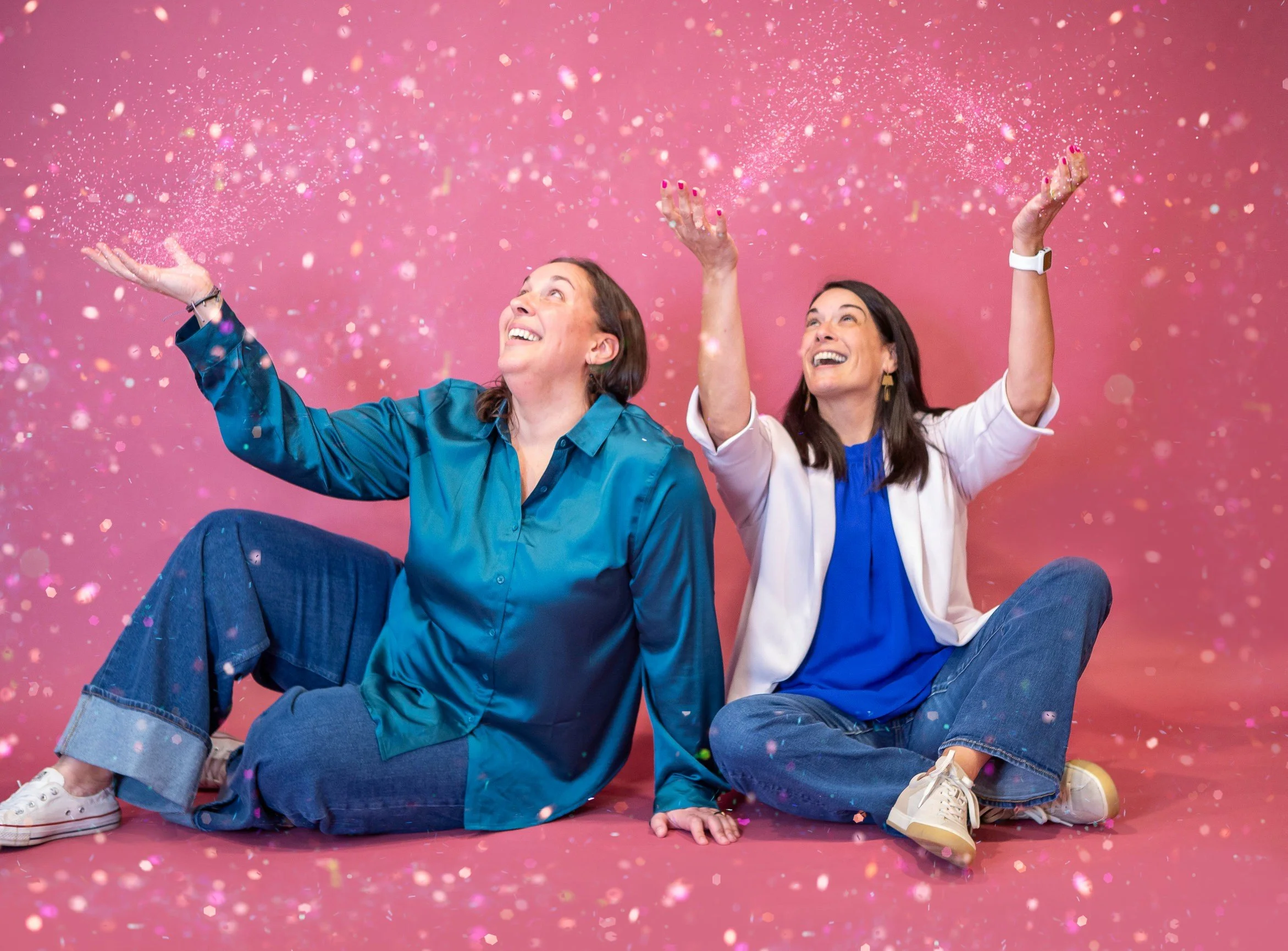 Two women sitting on the floor against a pink background, with colorful confetti falling around them. They are smiling and looking upward, appearing joyful and playful.