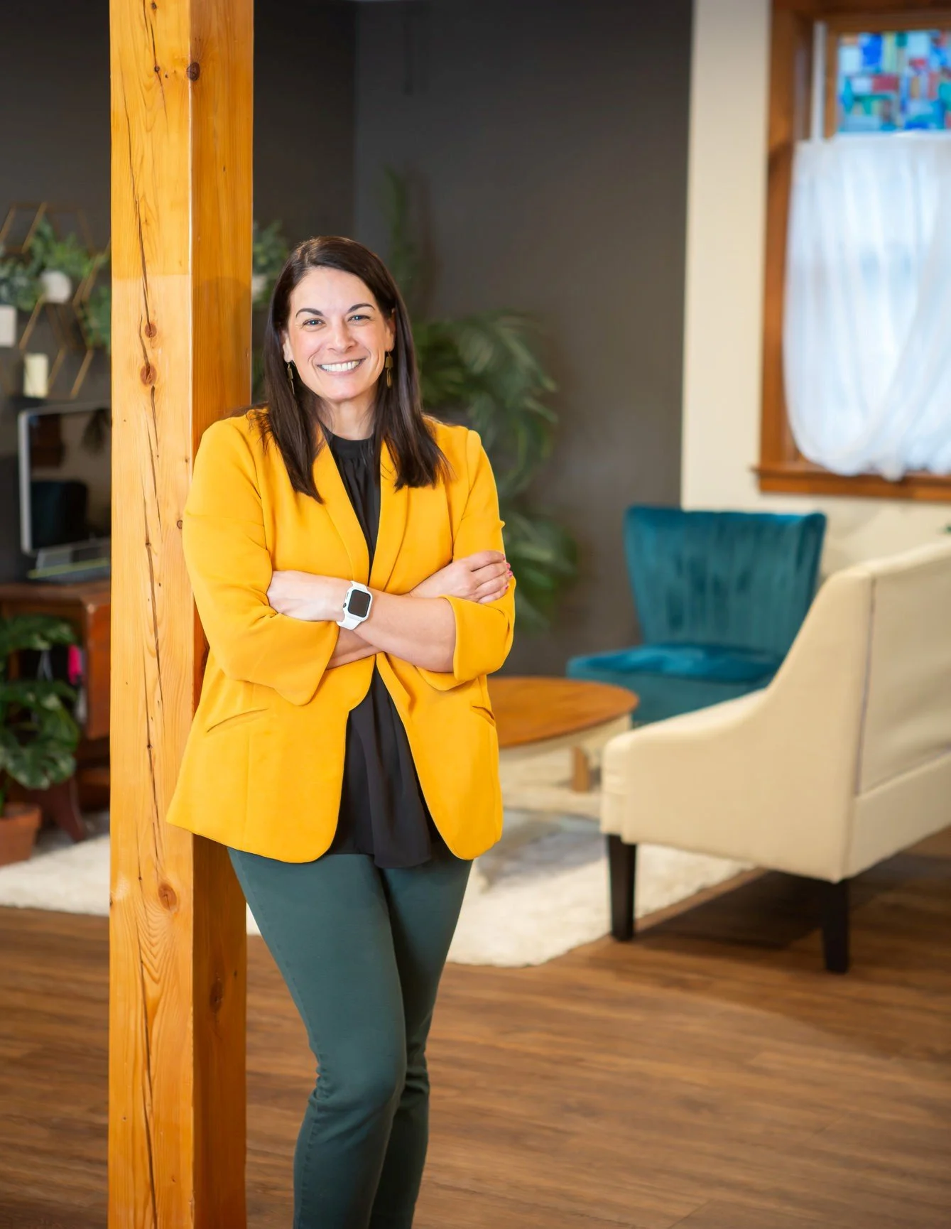 A woman with dark brown hair wearing a yellow blazer, black shirt, and teal pants, smiling with arms crossed, standing inside a cozy living room with a beige armchair, teal armchair, wooden coffee table, and houseplants.