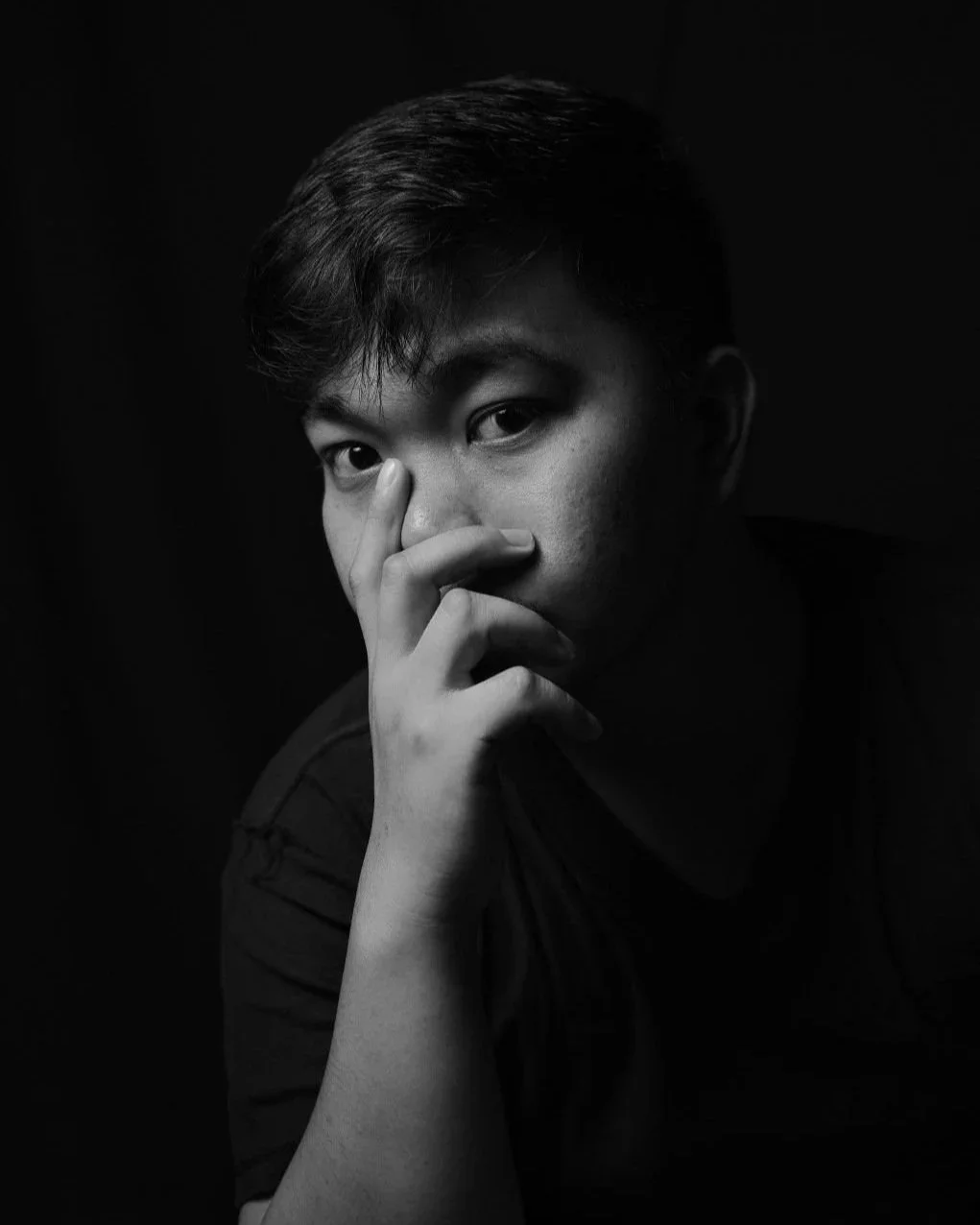 Black and white portrait of a young person with hand partially covering face, looking directly at the camera against a dark background.