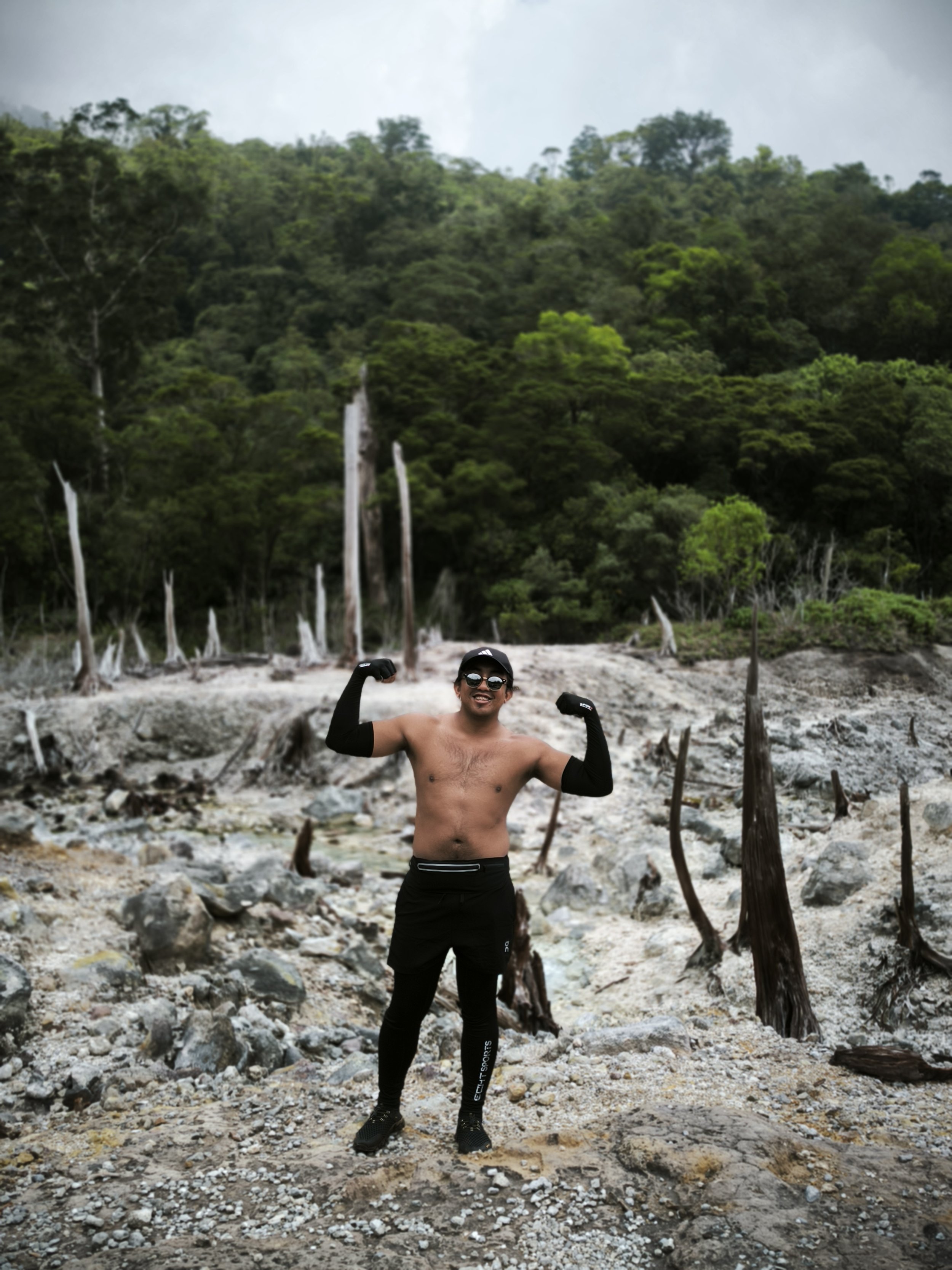 Shirtless man in black athletic clothing flexing in a rocky forest clearing