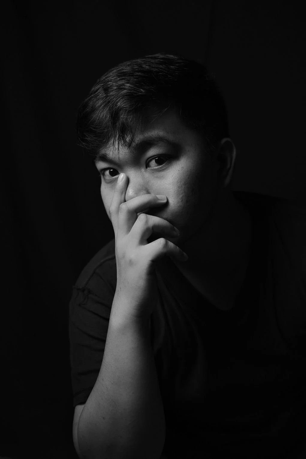Black and white portrait of a young man with short dark hair, looking intently at the camera, resting his chin on his hand, with a background that is dark and plain.