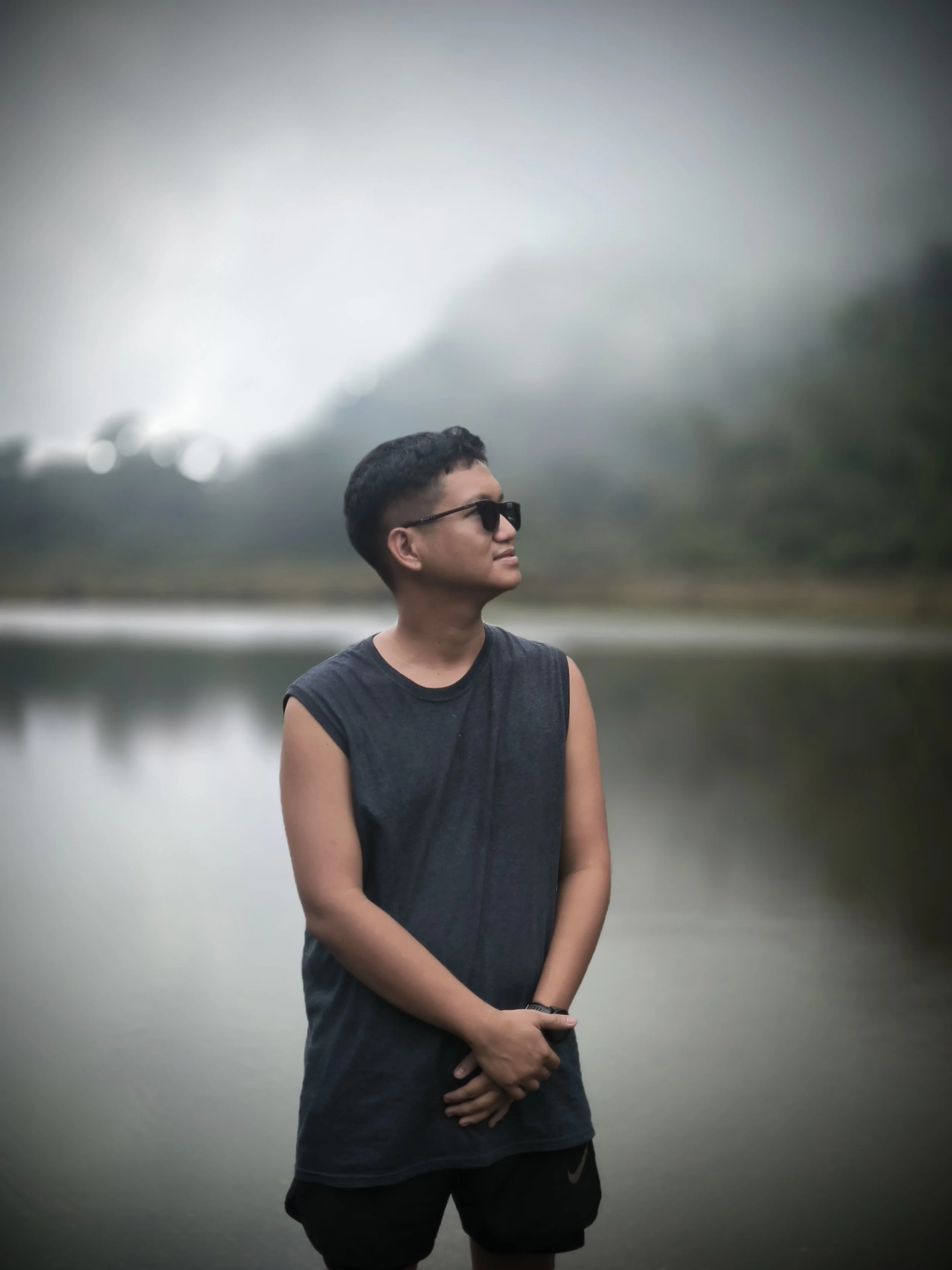 Person wearing sunglasses and a dark sleeveless shirt standing outdoors near a blurred body of water