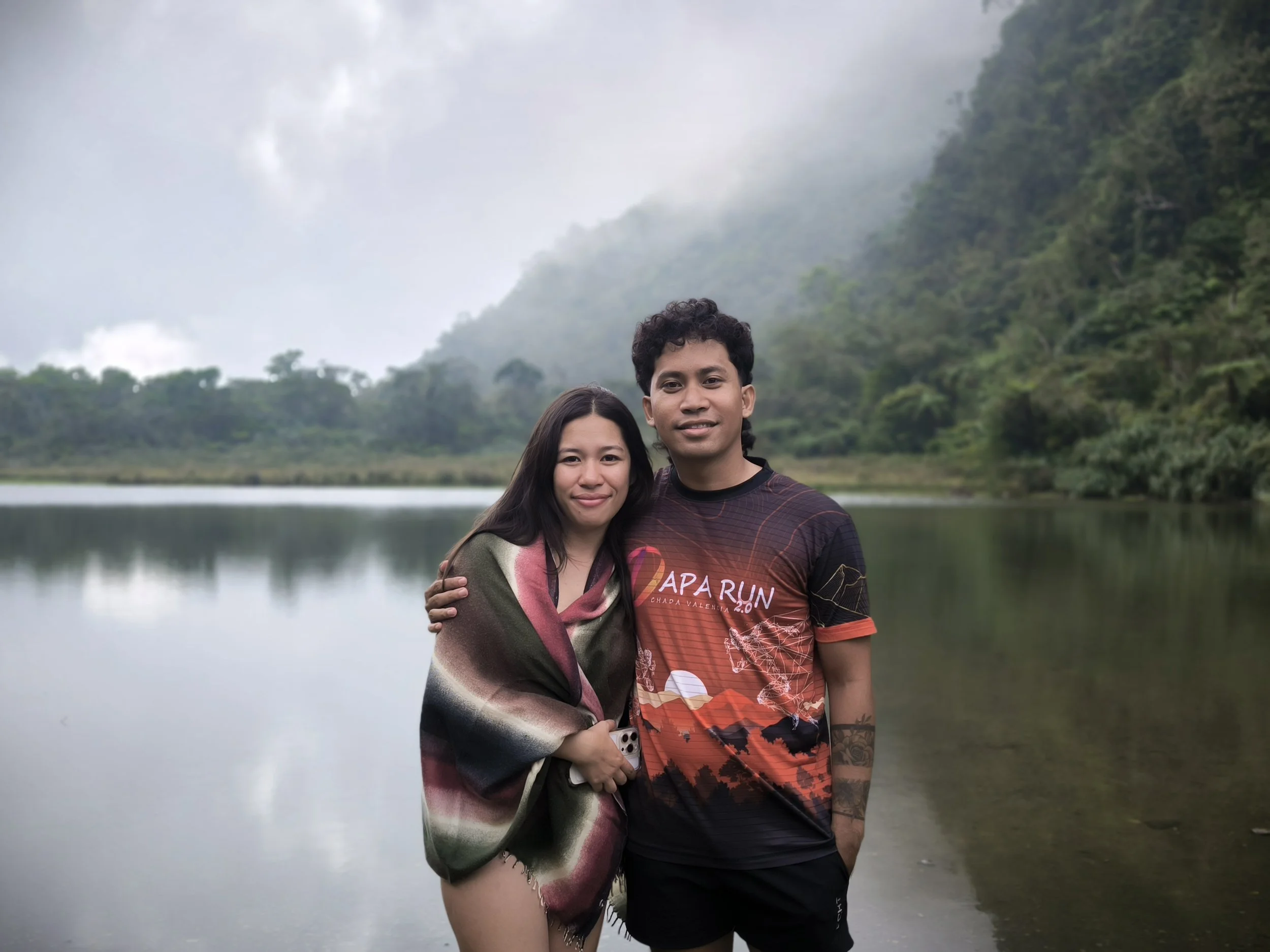 Couple posing beside a misty forest lake, with the man wearing a red sports jersey and the woman wrapped in a shawl.