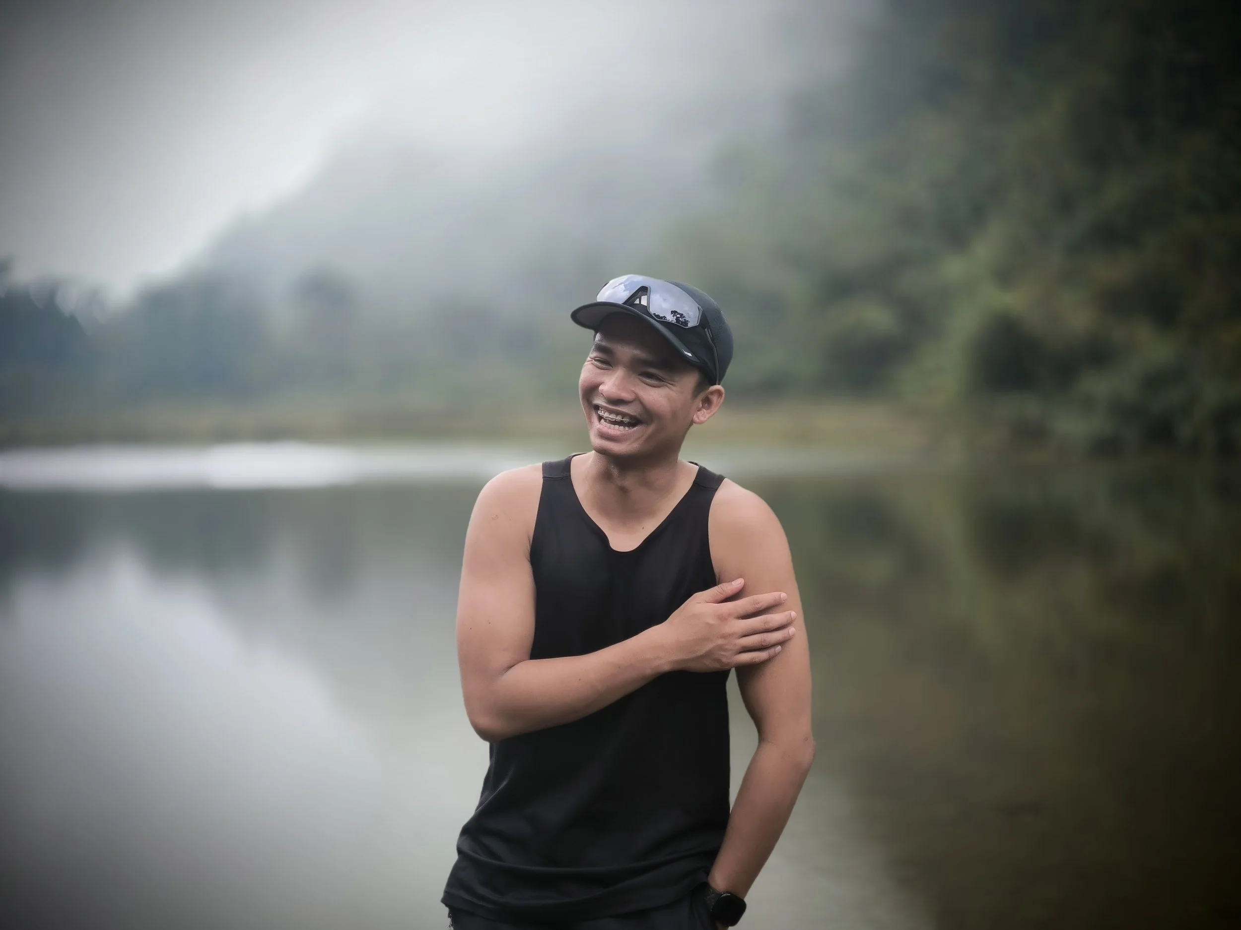 Smiling man in a black tank top and cap standing beside a misty lake surrounded by trees