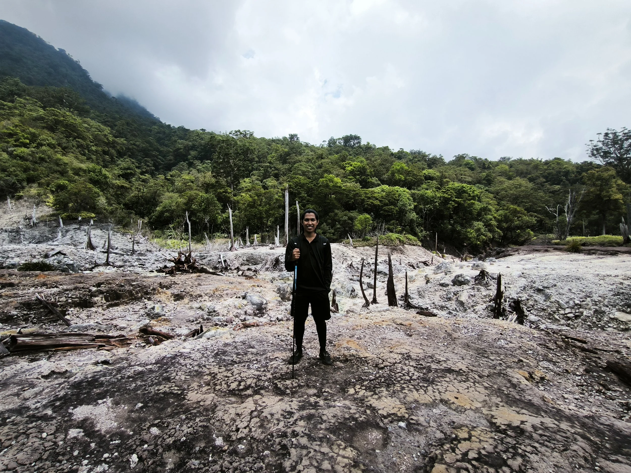 Hiker standing in a volcanic landscape with dead tree trunks, lush tropical forest, and a misty mountain behind him