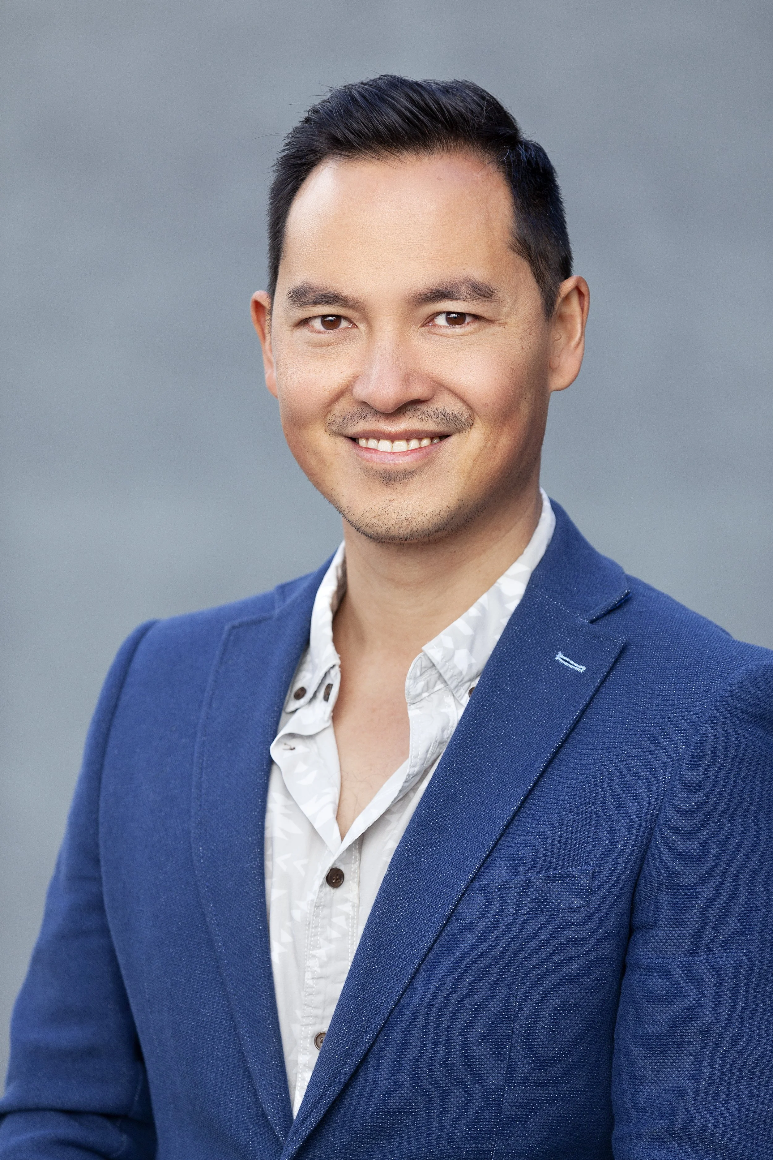 Headshot of a man with short black hair, smiling, wearing a blue blazer and a white shirt against a gray background.