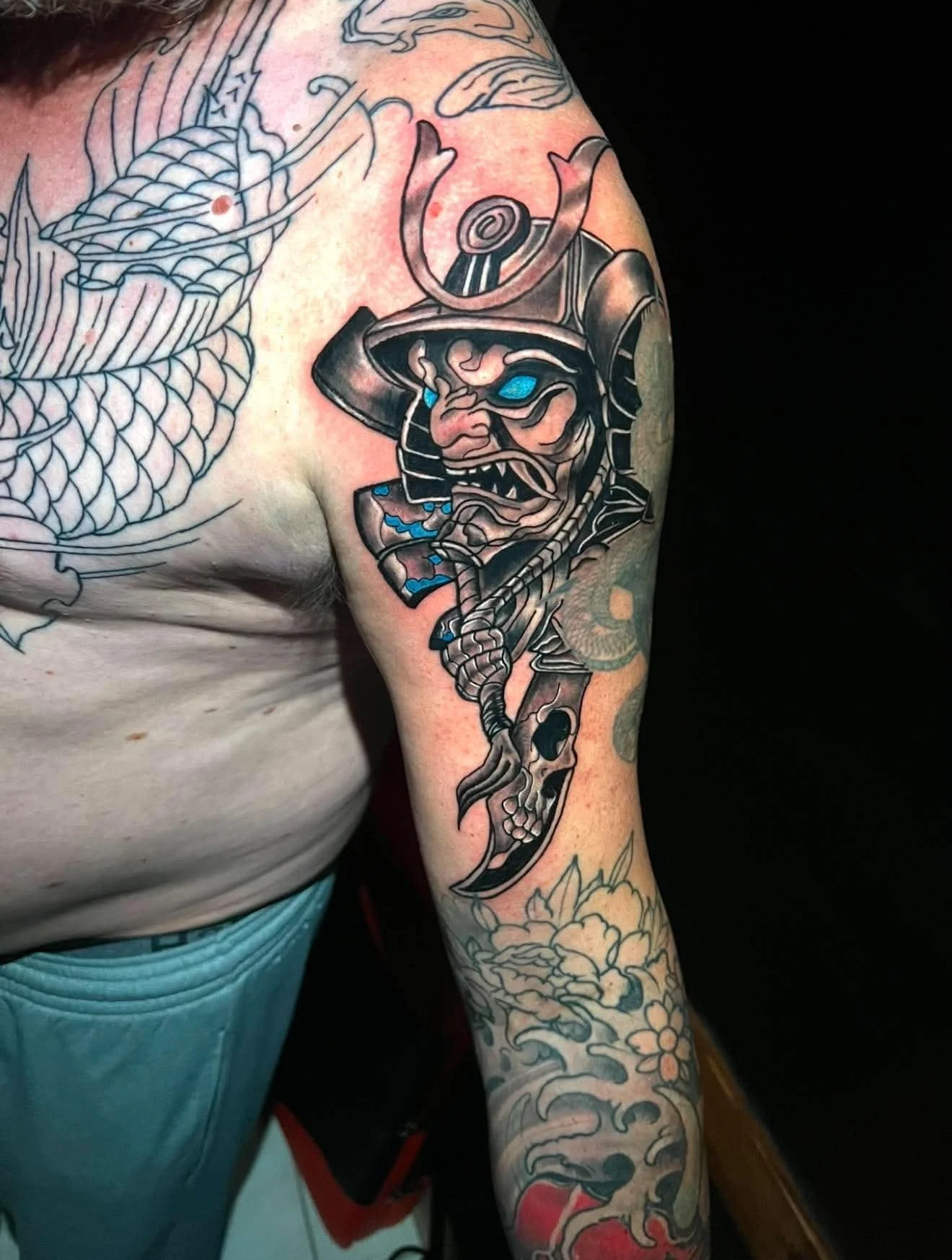 Tattoo of a fierce samurai warrior with blue eyes and traditional armor on a person's upper arm, surrounded by other tattoos including a koi fish and floral designs.