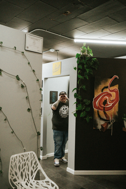 A person taking a mirror selfie in a modern interior space. The person is wearing a black t-shirt with a graphic print, blue jeans, and a cap. The room has decorative string lights with greenery, a white chair with a geometric design, and artwork on the walls, including an abstract painting with warm colors.