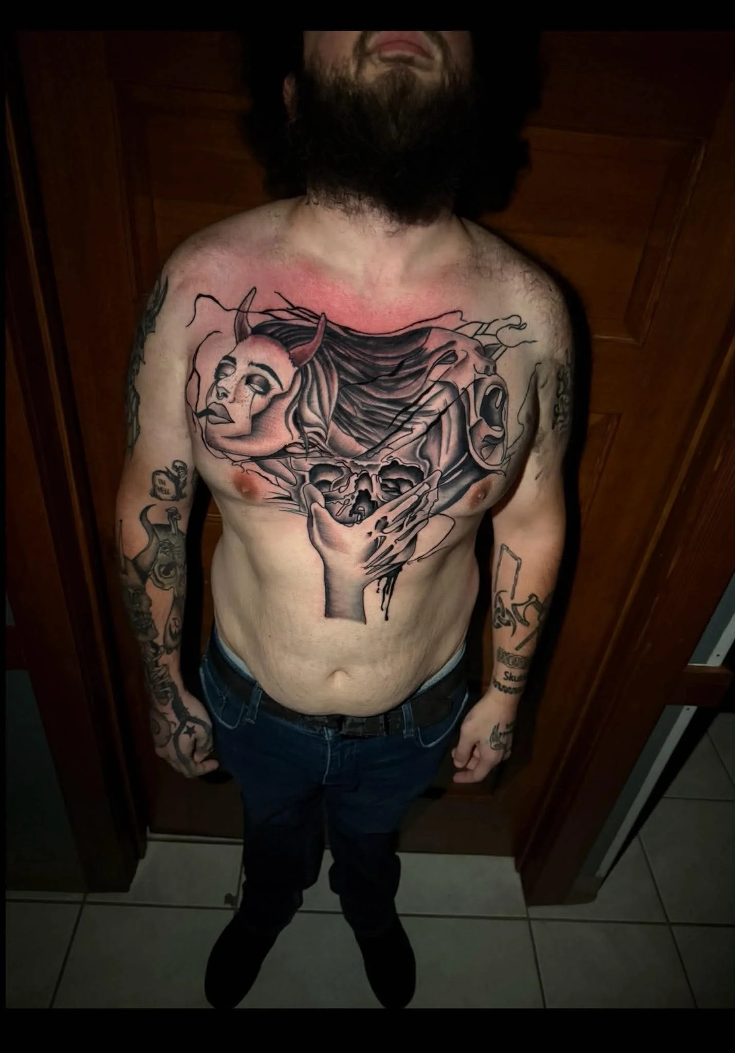 A shirtless man with tattoos on his arms and chest, standing in a room with wooden cabinets, showing a large tattoo on his chest of a woman with long hair, horns, and a sad expression.