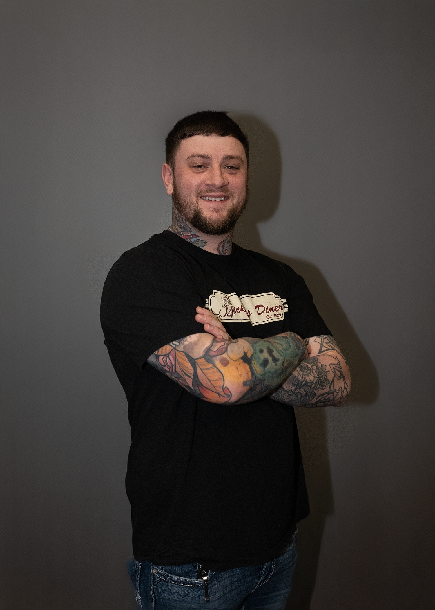 A man with tattooed arms, a beard, and short dark hair stands against a plain dark background, smiling and crossing his arms. He is wearing a black T-shirt with a graphic logo and design on the front.