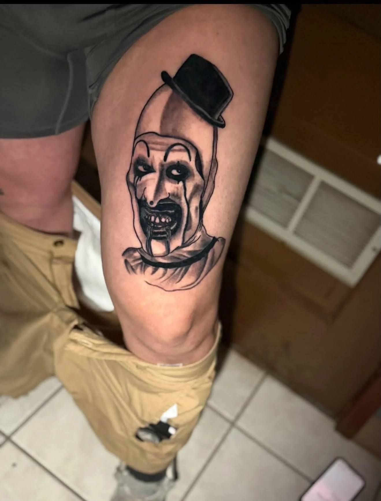 Tattoo of a clown with dark makeup and teeth, holding a small black object, on a person's upper arm.