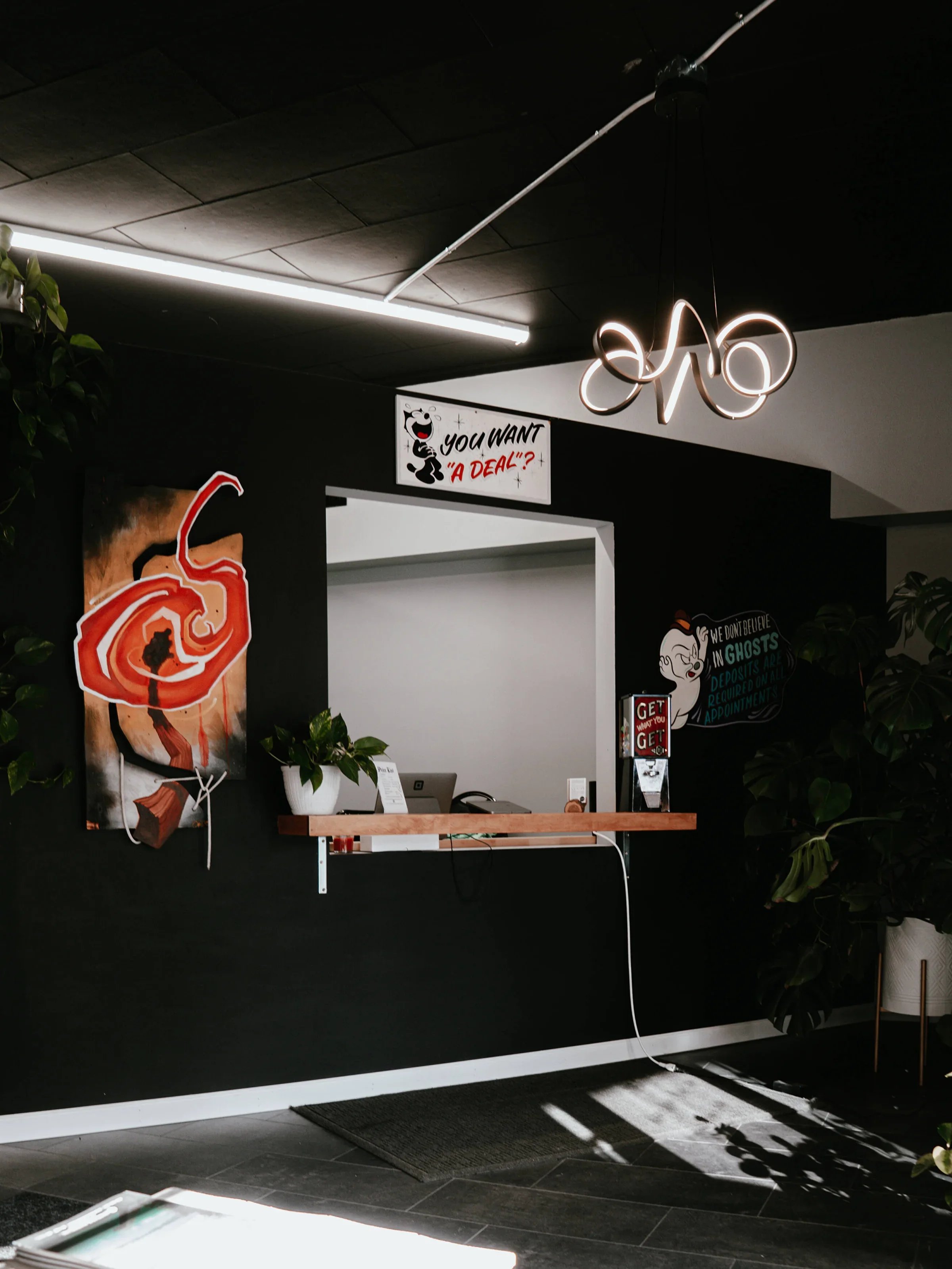 Indoor scene of a reception area with a black wall featuring colorful artwork, a small wooden counter, and decorative wall signs, including a cartoon panda with a speech bubble that says 'You want a deal?'. There are green plants and a modern light fixture hanging from the ceiling, with sunlight casting shadows on the floor.