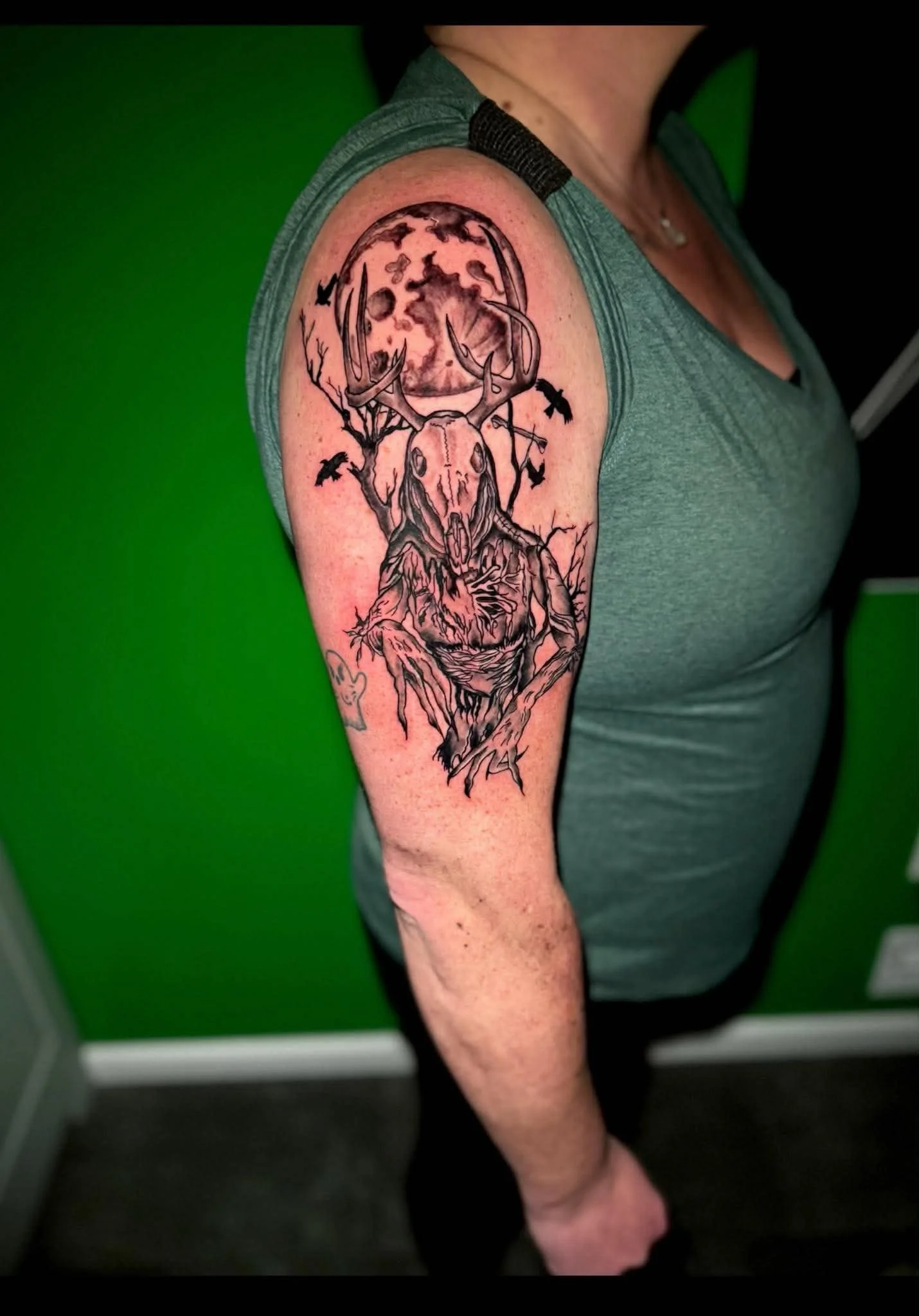 Tattoo of a deer skull with antlers, set against a moon and tree branches on a person's upper arm.