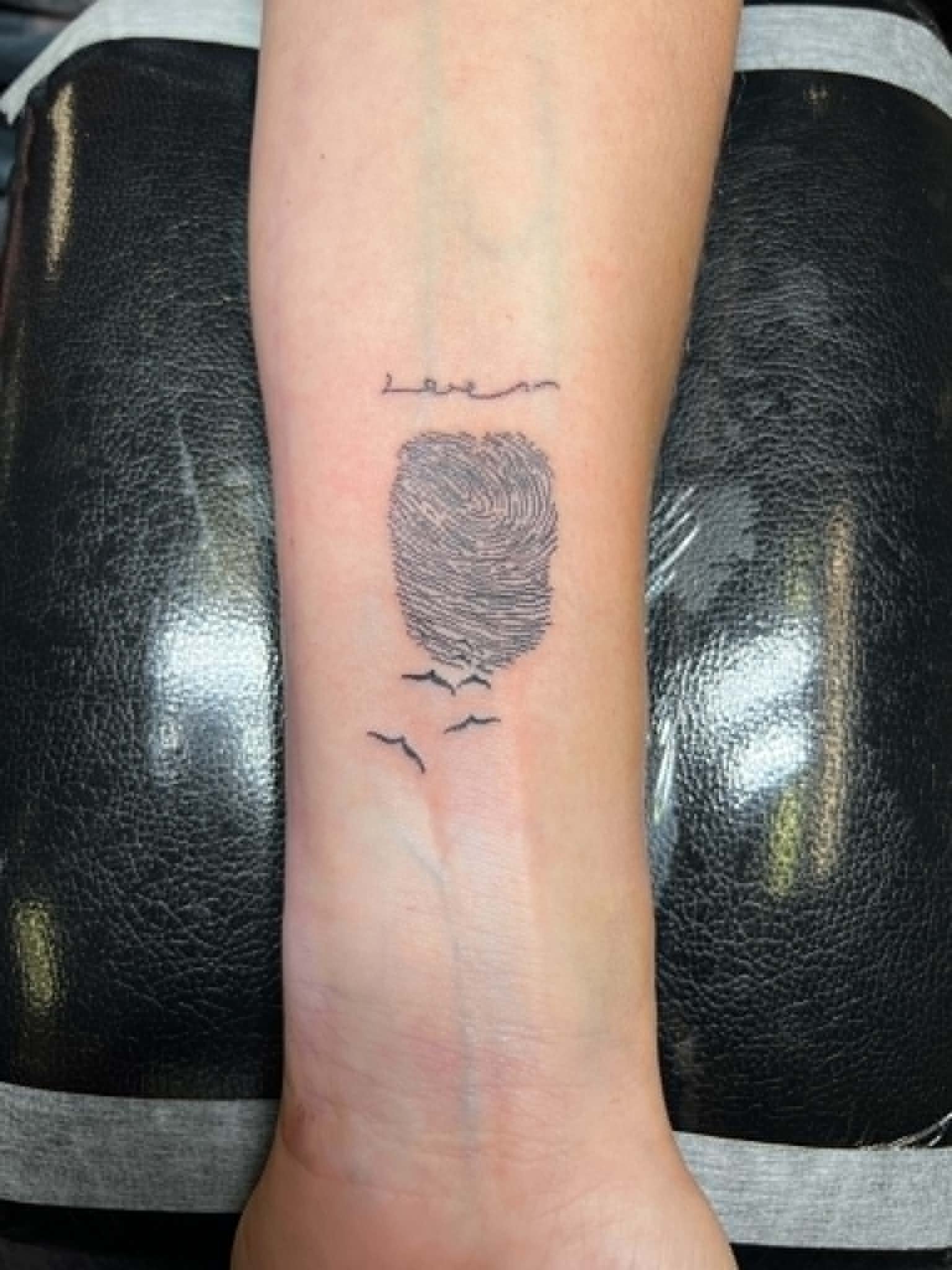 Tattoo of a fingerprint with small birds flying below and a written word above on a person's inner forearm