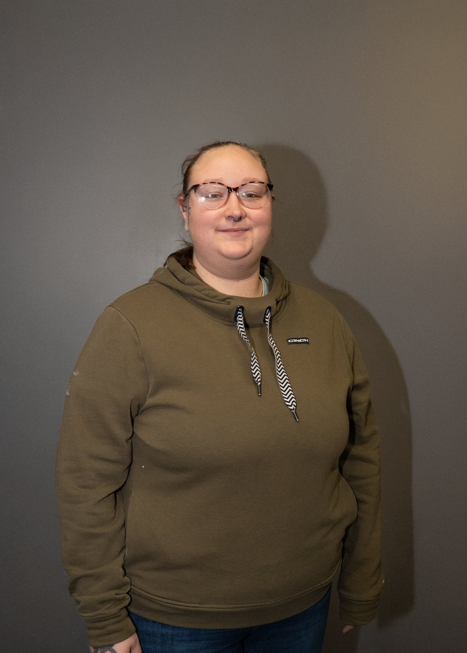 A person with glasses, a septum piercing, and a tattoo on their hand, wearing a brown hoodie with drawstrings and dark jeans, standing against a plain gray wall.