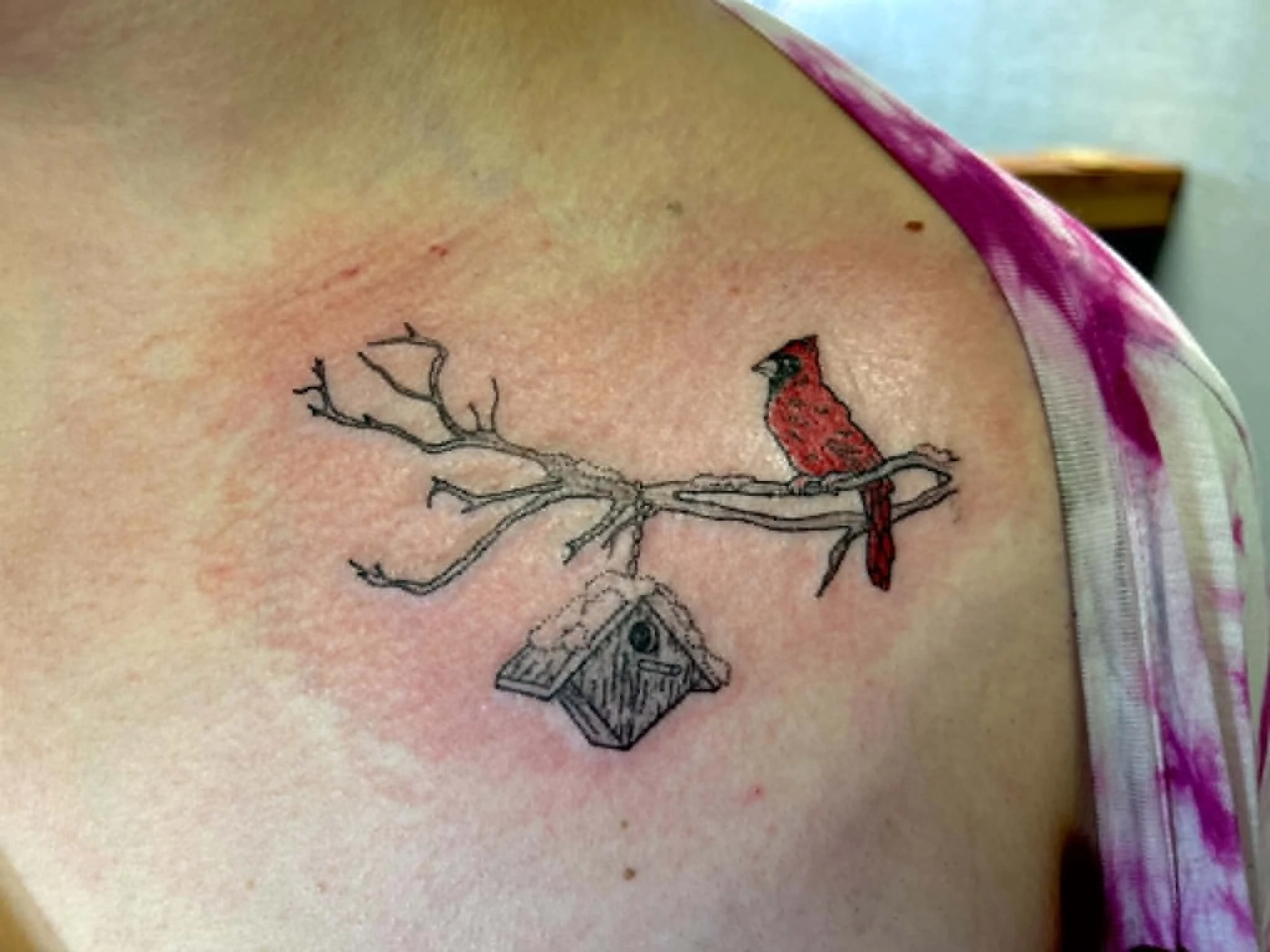 A tattoo of a bird sitting on a tree branch. The bird is red with black eyes and beak, and the branch is detailed with twigs. The tattoo is on someone's skin, with a pink shirt collar visible at the top edge.