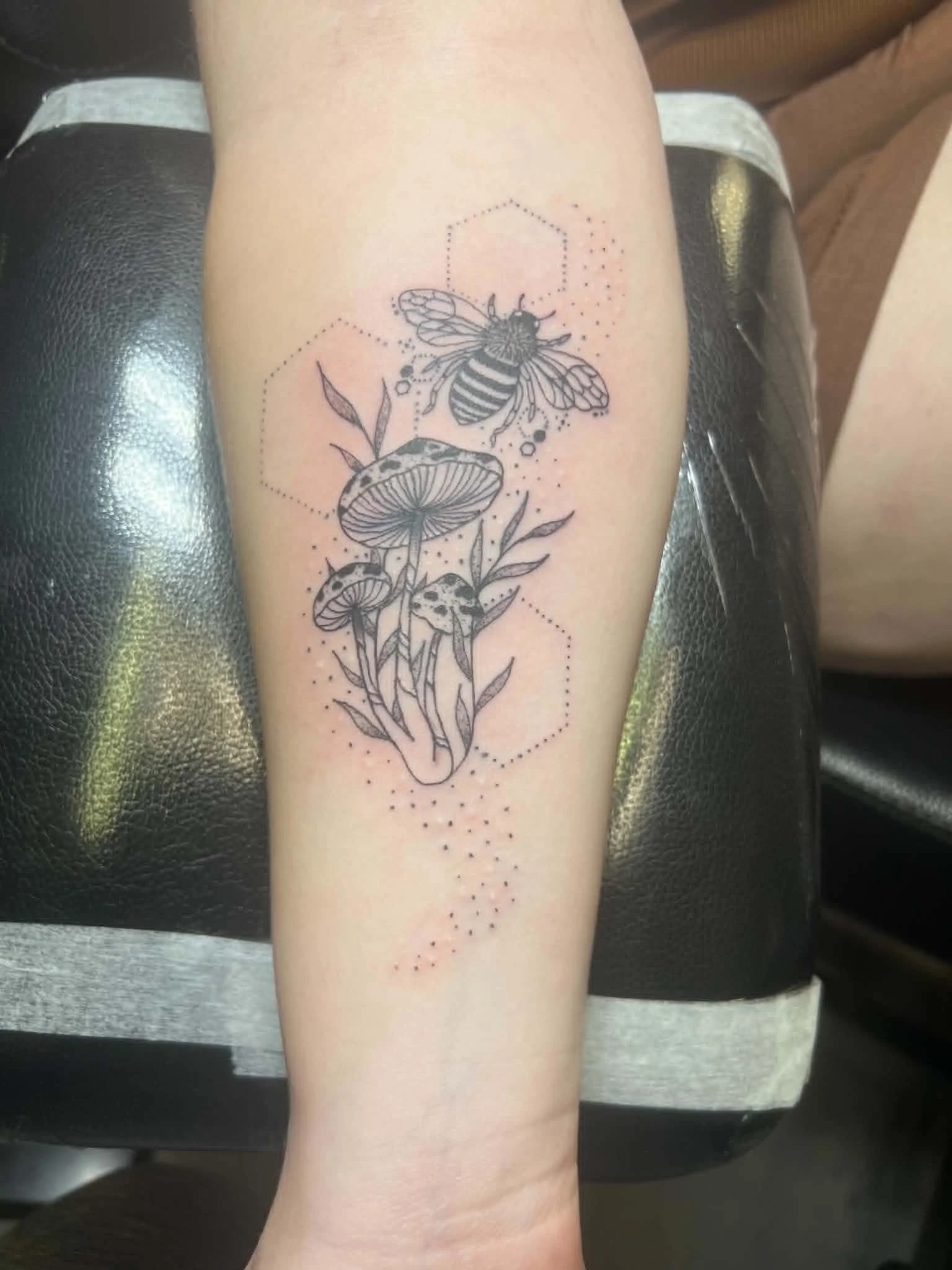 Tattoo of a bee, mushrooms, and foliage on a person's arm, with geometric dotted lines around the design.