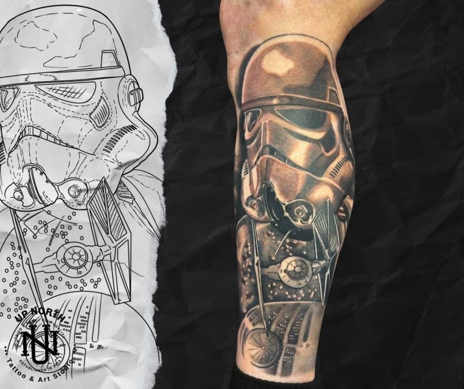 A detailed black and gray tattoo of a Stormtrooper helmet from Star Wars on a person's forearm, next to the tattoo's design sketch.