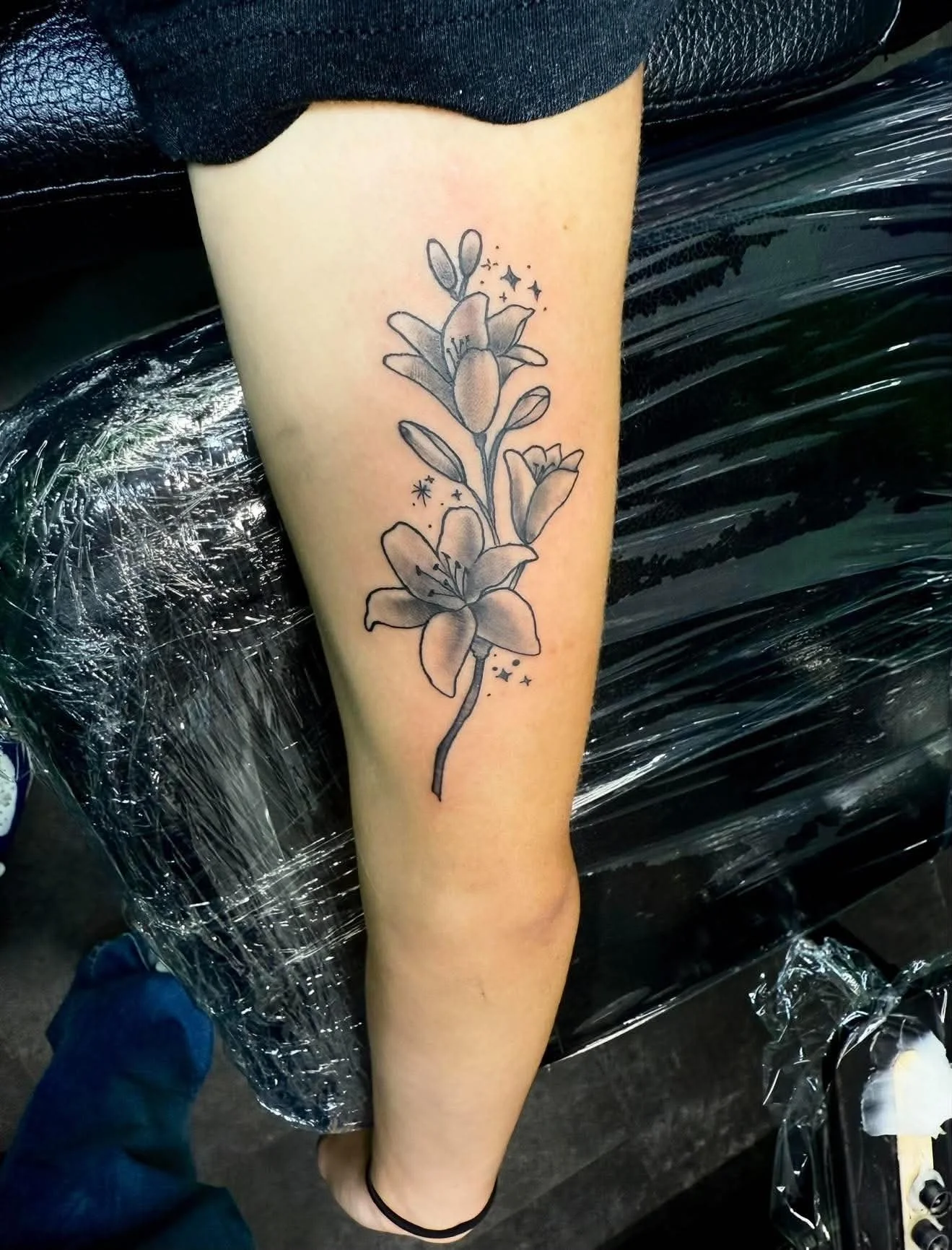 A black and gray floral tattoo on a person's forearm, depicting a branch of flowers with small stars and sparkles around it.