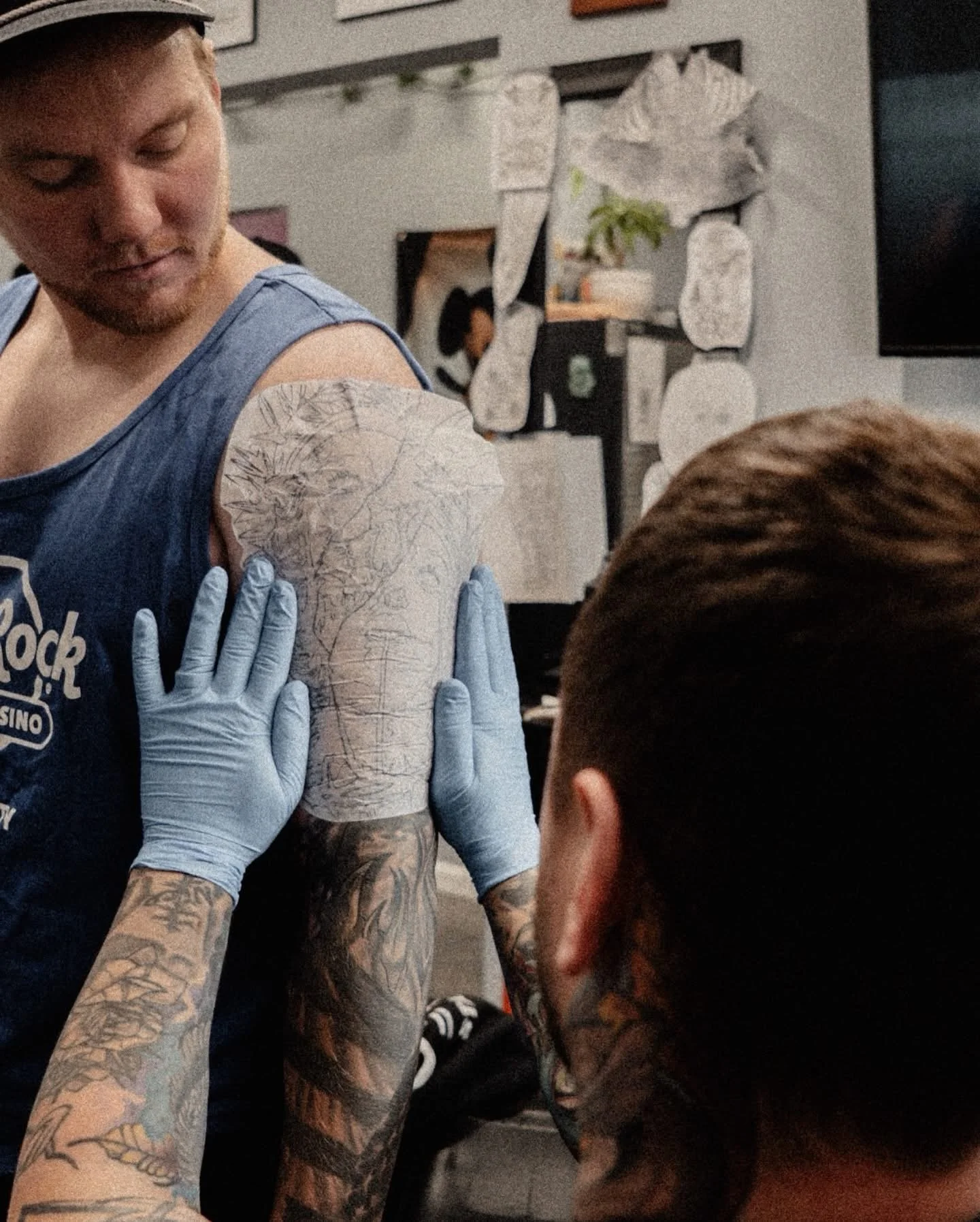 A person with extensive tattoos receiving a vaccination injection in their upper arm from a medical professional wearing gloves. The background shows a wall with decorative stones and a small plant.