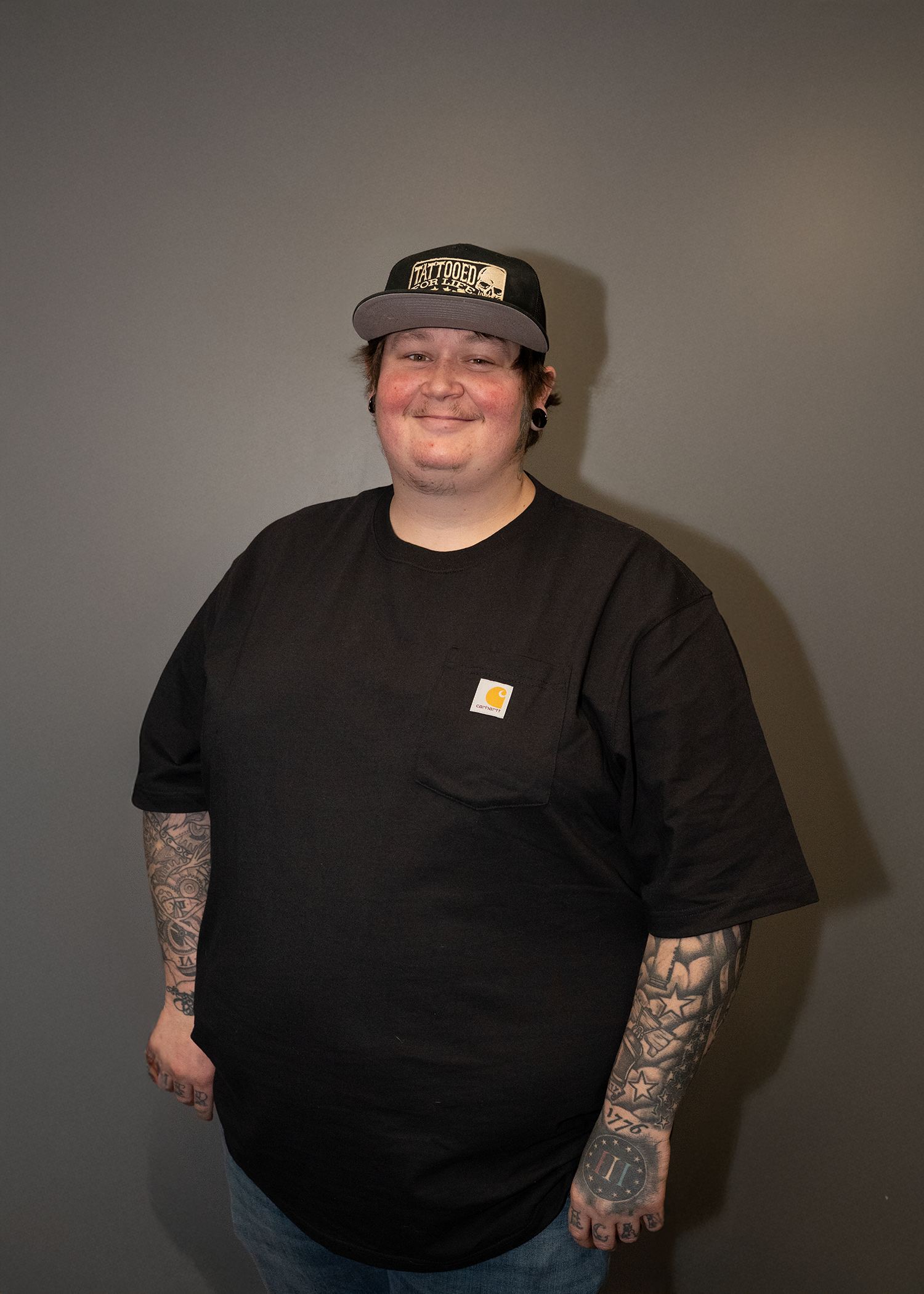 A young man with multiple tattoos on his arms, wearing a black Carhartt T-shirt, blue jeans, and a black and gray baseball cap with a skull and the words 'Tattooed for Life' on it, standing against a plain gray wall.