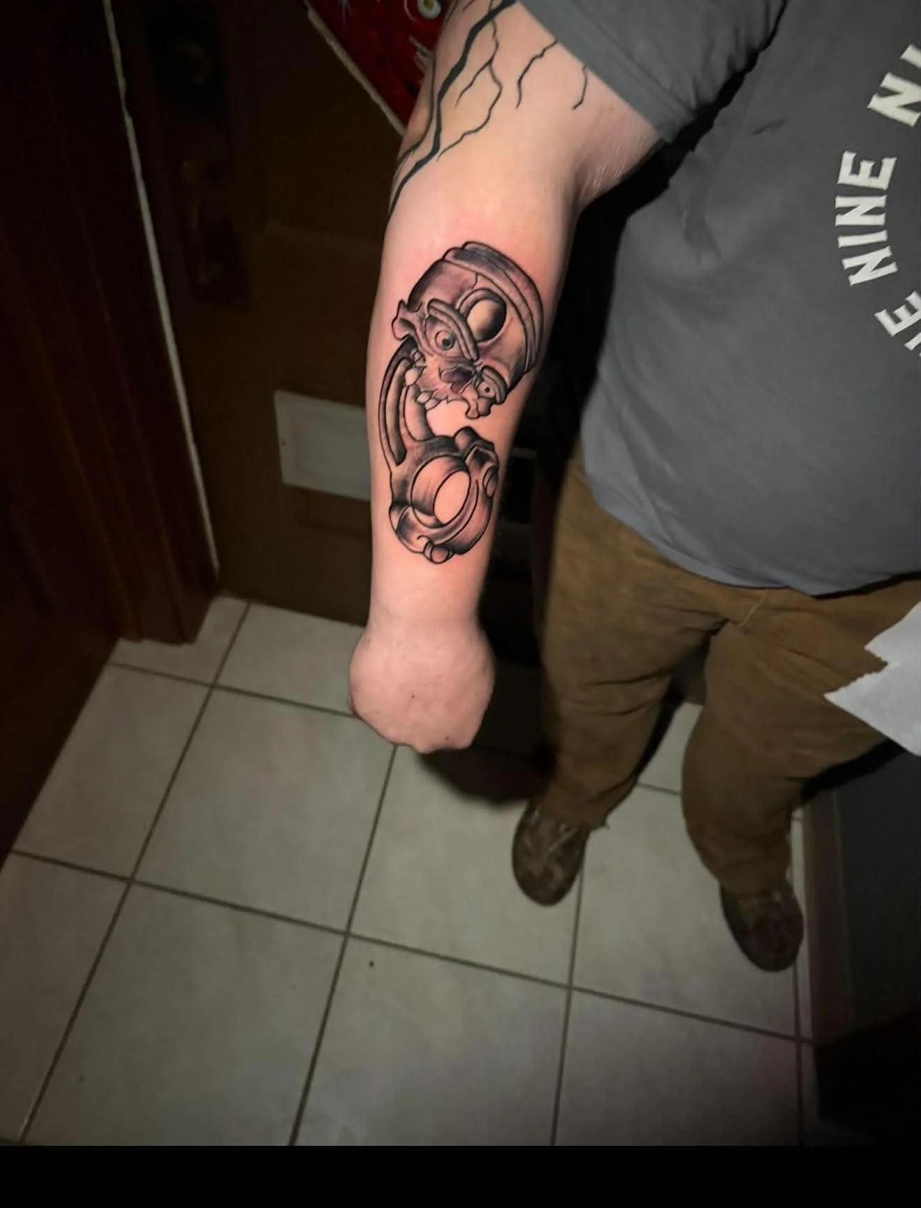 A person's arm with a detailed black and gray tattoo of a revolver with a cartoonish skull inside the barrel, on a tiled floor background.